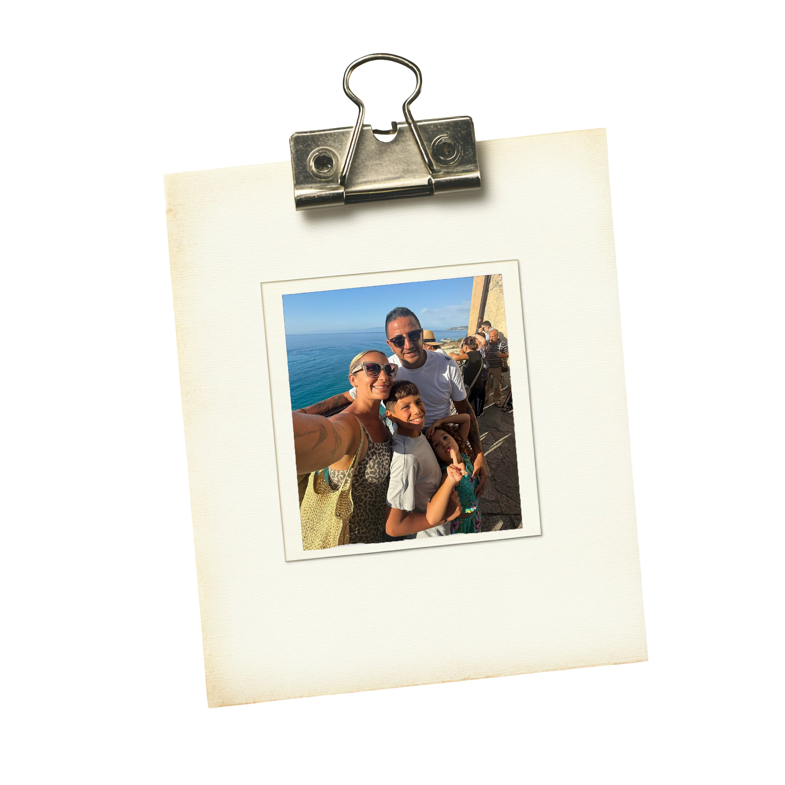 A printed photograph held with a binder clip, showing smiling adults and children outdoors near a body of water, under a clear blue sky.