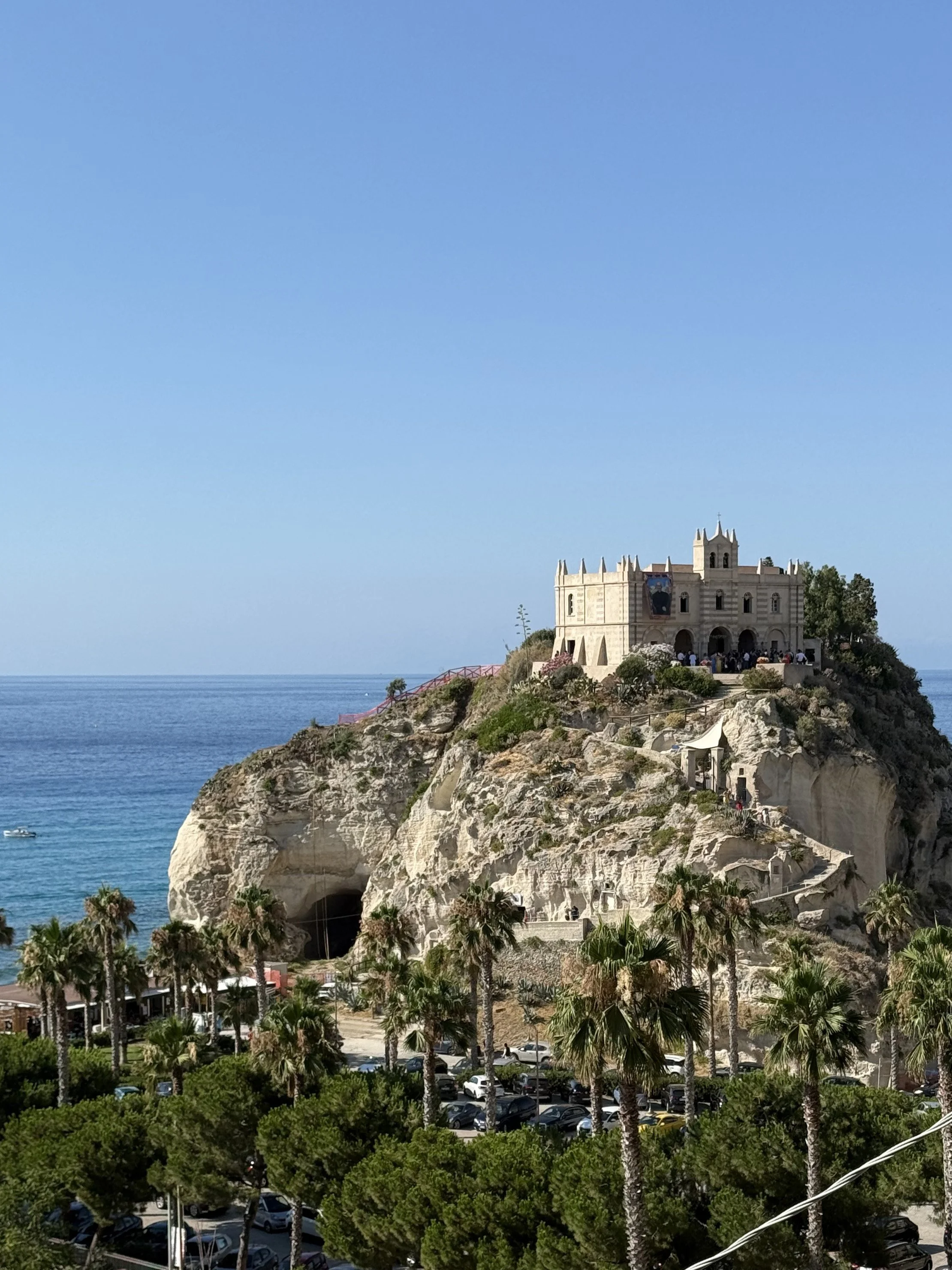 View of a historic castle-like building on a rocky hill overlooking the ocean, with palm trees and a parking lot in the foreground on a clear day.