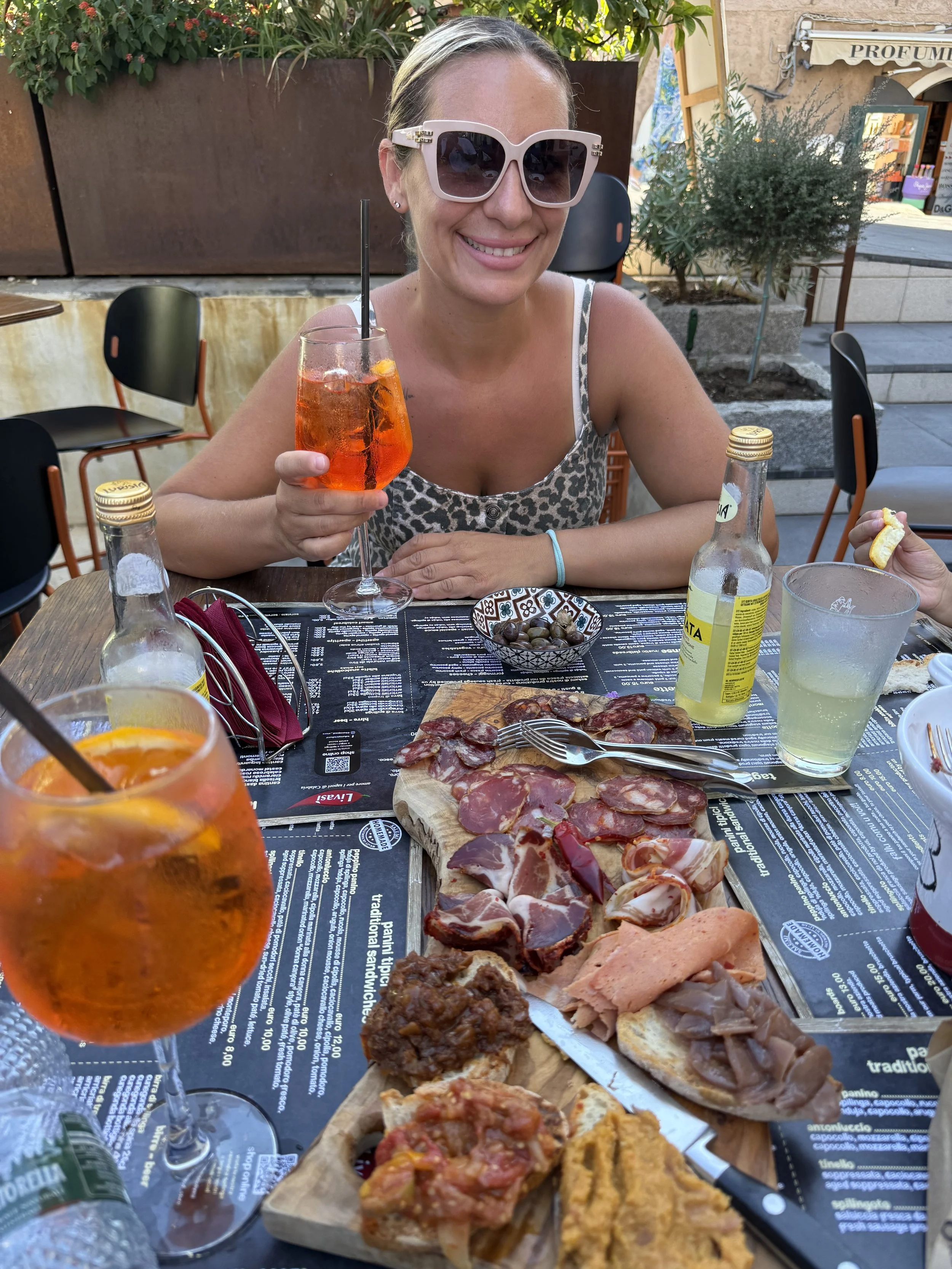 A woman wearing large sunglasses and a patterned top sitting at an outdoor table smiling while holding a glass of orange-colored drink with ice and a black straw. The table has a variety of foods including a charcuterie board with sliced meats and toppings, bottles of water and soda, and a small bowl of marbles. In the background, there are chairs, plants, and a building with a sign that reads 'PROFUMERIA'.