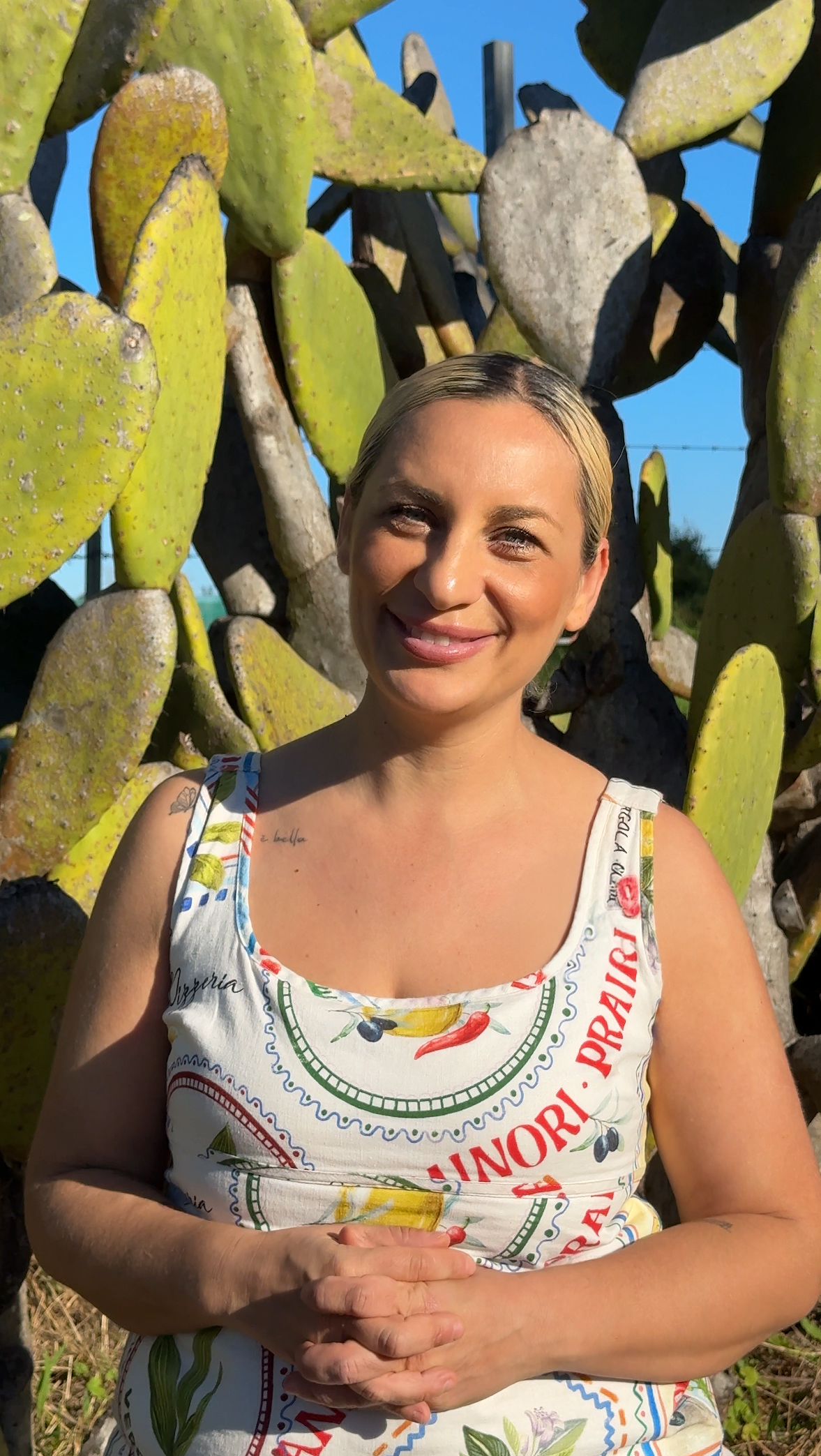 A woman with short blonde hair smiling outdoors, standing in front of large prickly pear cactus pads, under a clear blue sky.