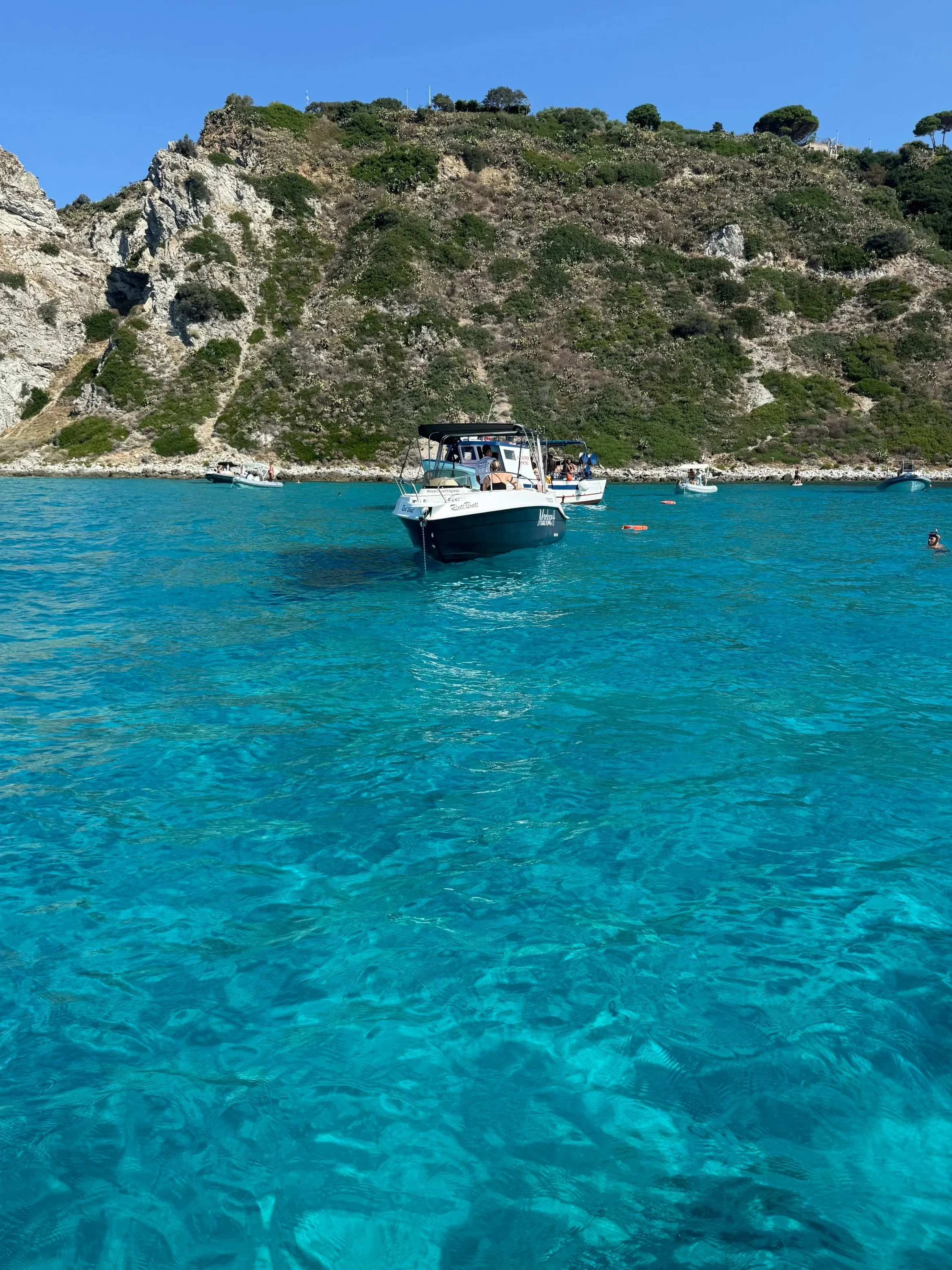 Boats floating on bright turquoise water near a rocky hillside covered in green vegetation.