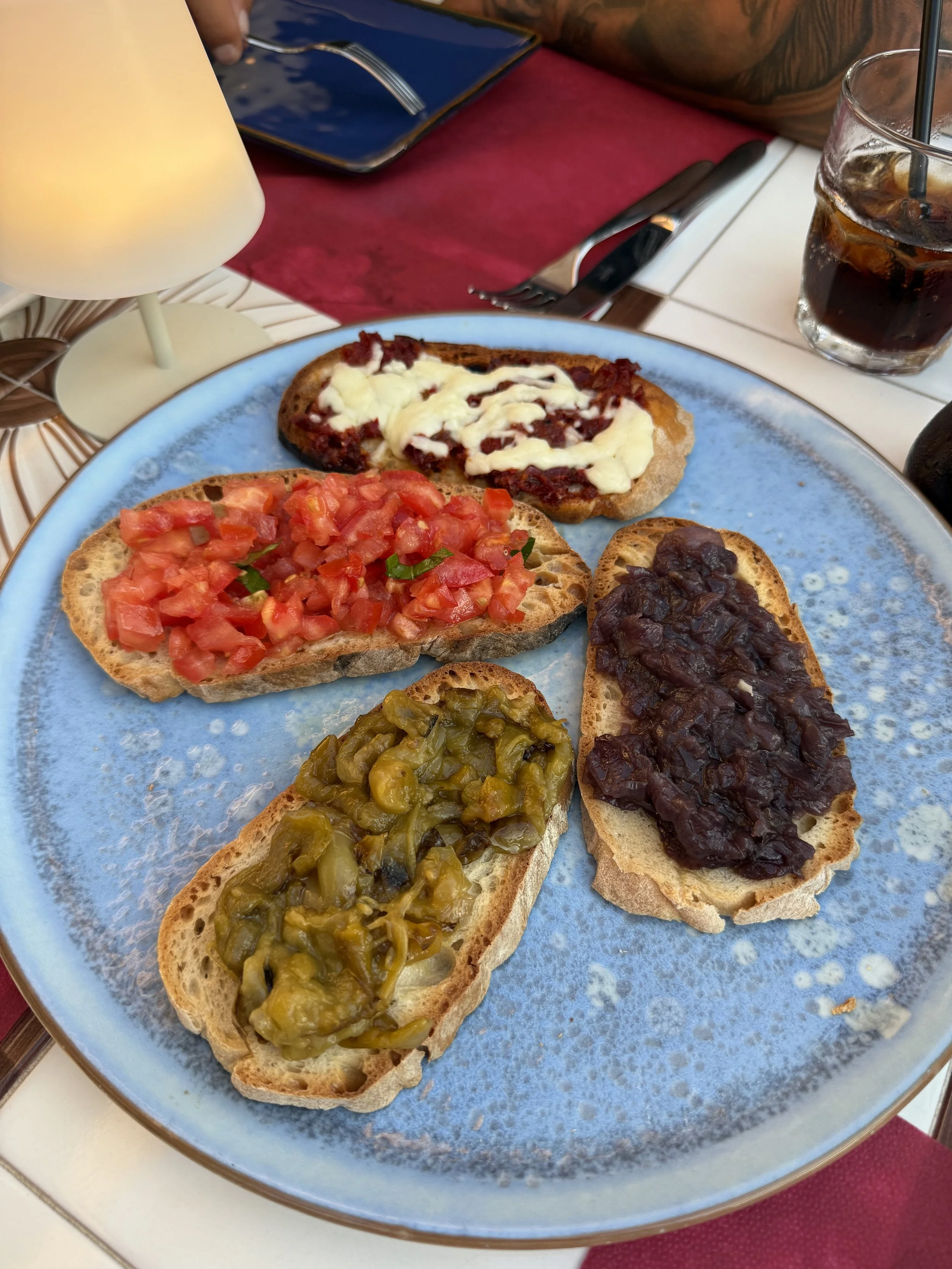 Plate with five slices of toasted bread topped with various spreads, including tomato and green pepper, black bean, olive tapenade, cheese, and fruit jam. A glass of cola with ice and a straw is beside the plate.