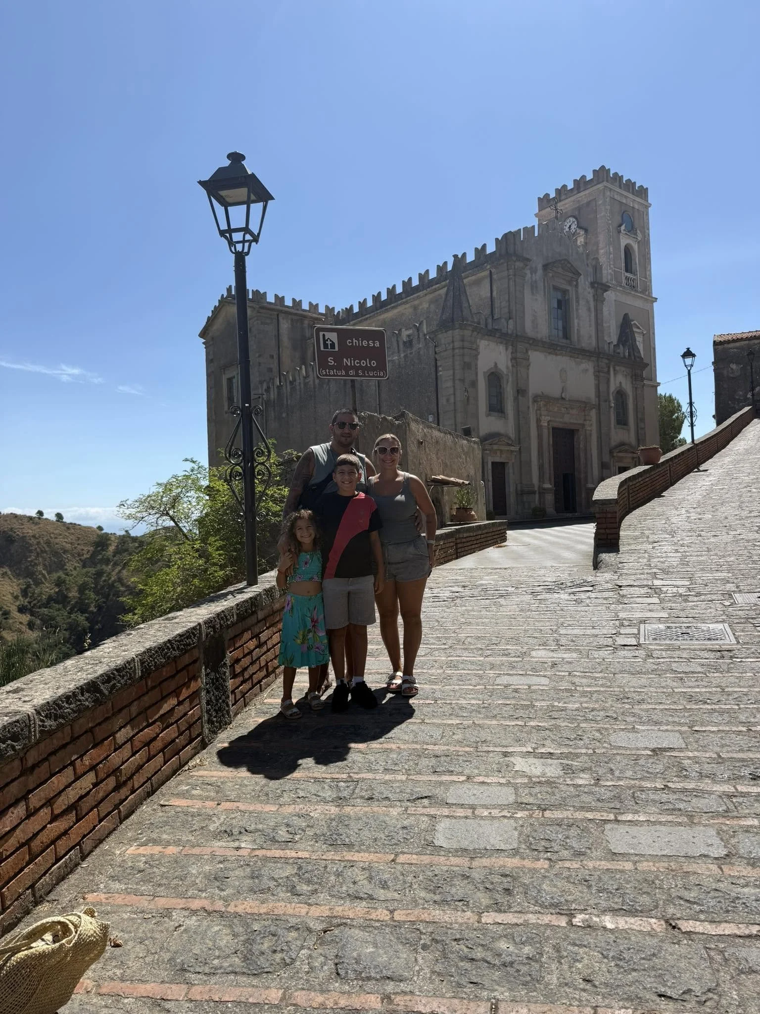 A family of four standing on a cobblestone street in front of a historic church with a clock tower, clear blue sky above, and scenic hilly landscape in the background.