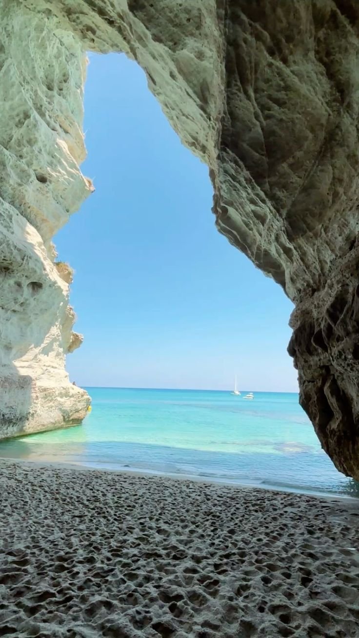 A view of a beach seen through a rocky cave opening, with turquoise water and sailboats in the distance under a clear blue sky.