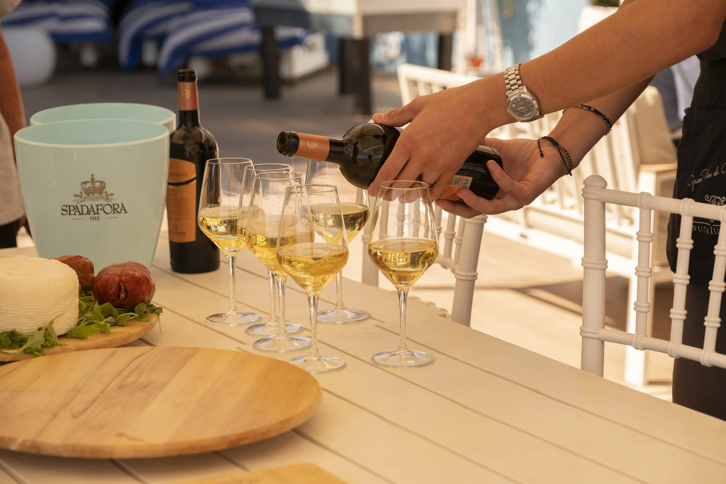 A person pouring wine into glasses on a table set for a meal, with cheese, apples, and a wine bucket nearby, outdoor setting with blurred chairs and water in background.