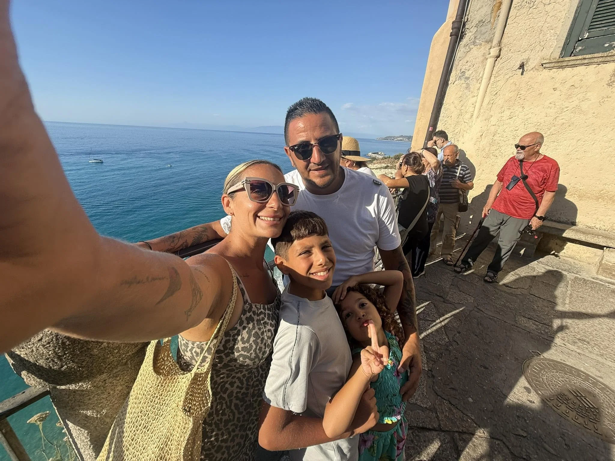 A group of people, including children and adults, taking a selfie together outdoors near a body of water, with a stone building and a blue sky in the background.