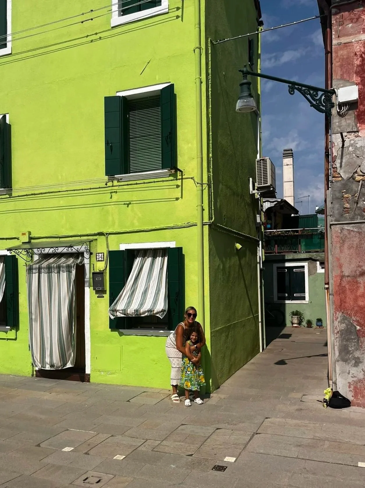 A woman and a young girl standing in front of a lime green building with green shutters and striped curtains, on a sunny day. The woman is wearing sunglasses and a sleeveless top, while the girl is in a brightly colored dress.