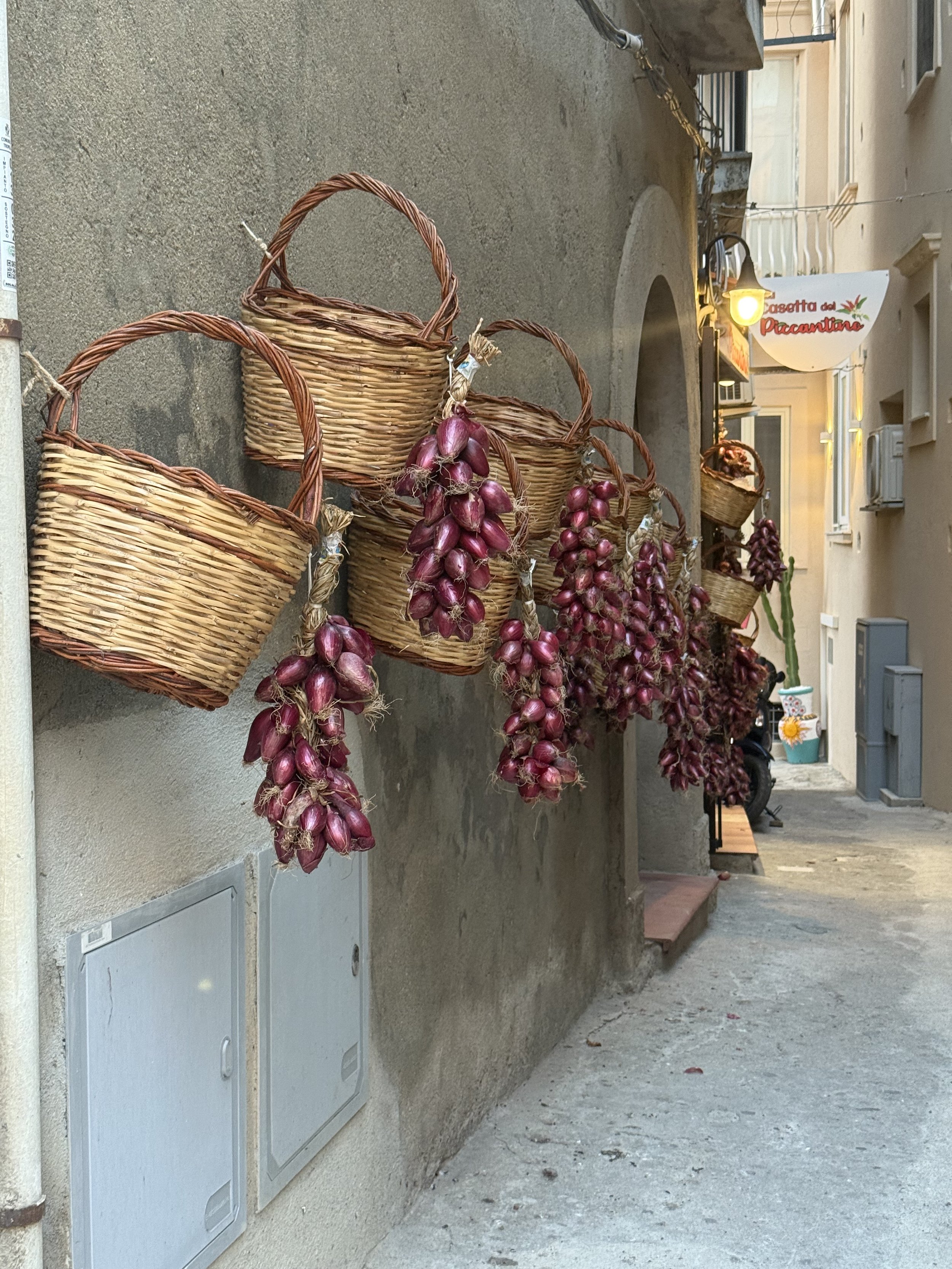 Several bunches of red onions hanging on wicker baskets attached to a wall in a narrow street.