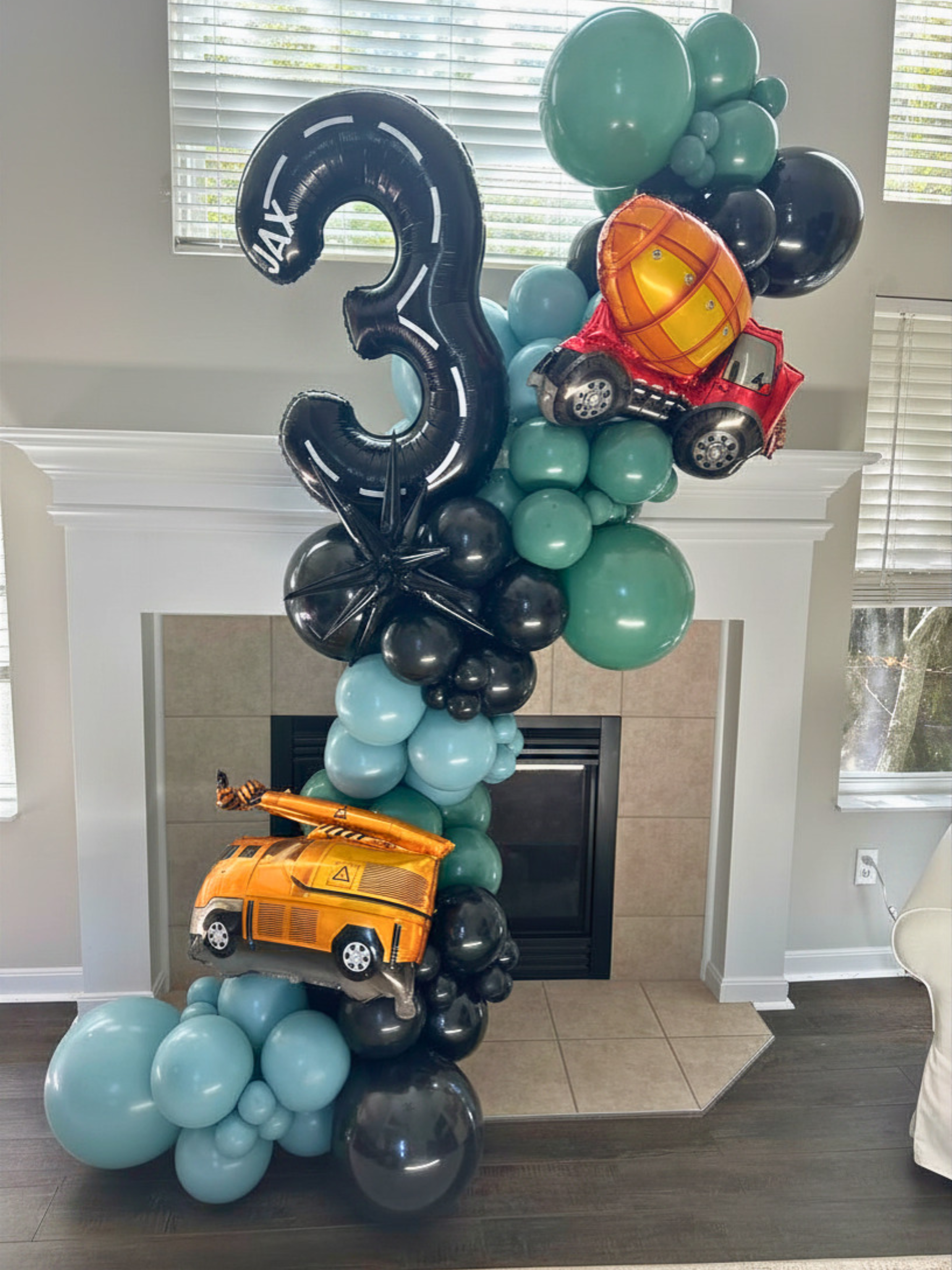 Balloon arrangement with a large black number 3 balloon, a traffic light-shaped balloon, action figure balloons, and cluster of black, teal, and blue balloons, set up in front of a fireplace and window.