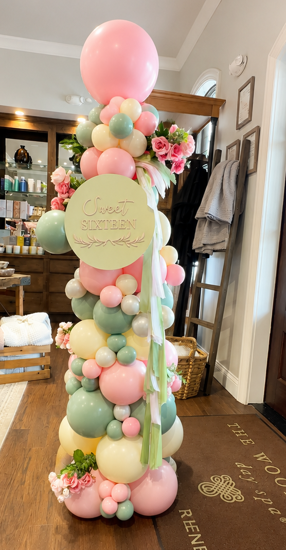 A tall balloon tower featuring pastel pink, green, yellow, and cream balloons, decorated with pink roses, a round sign that says 'Sweet Sixteen,' and green, pink, and white ribbons. It is set up indoors on a wooden floor near a doorway and some shelves.
