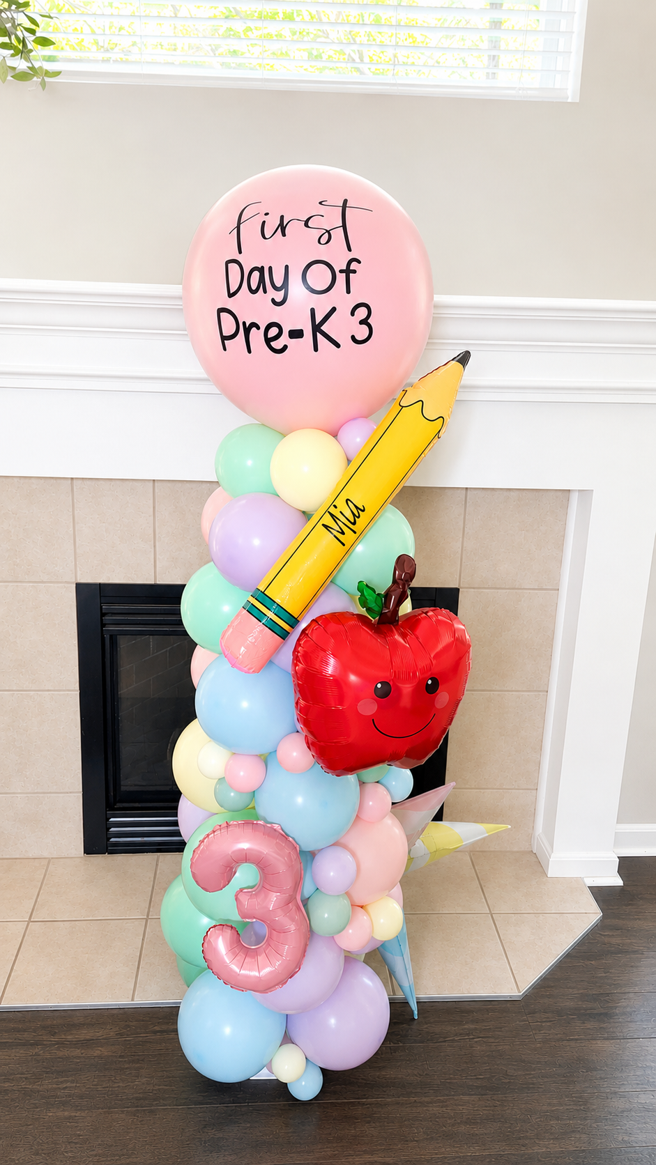 Colorful balloon arrangement for a child's first day of pre-kindergarten, featuring a pink balloon with black writing, a large yellow banana-shaped balloon with the name Mia, a red apple-shaped balloon with a smiling face, and a pink number three balloon, all set against a background of a fireplace and white window blinds.