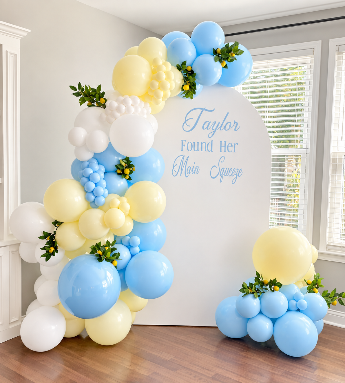 Balloon arch with yellow, white, and blue balloons, decorated with green leaves and small yellow fruits, adjacent to a white backdrop with blue cursive and print text that reads, "Taylor Found Her Main Squeeze."