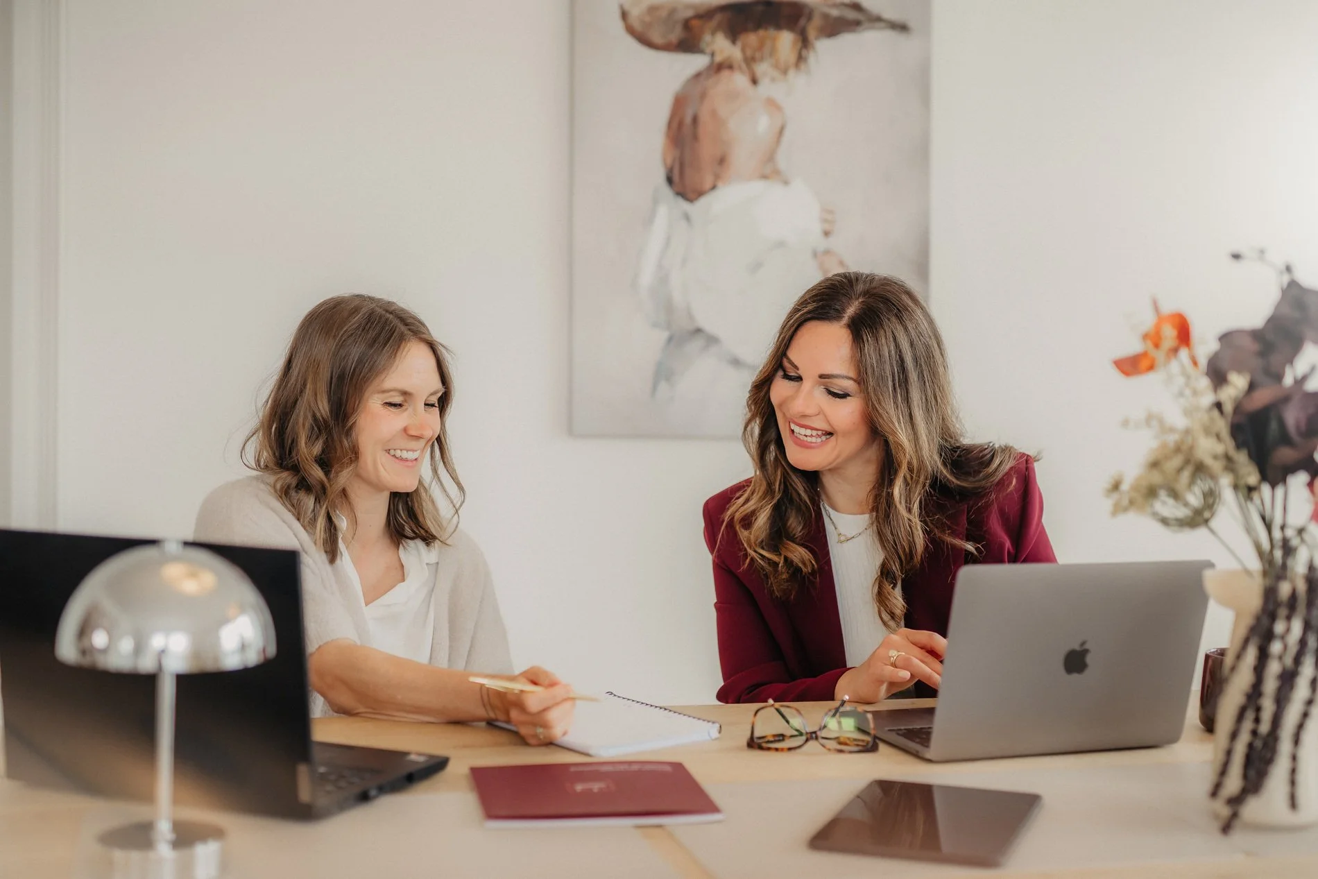 Zwei Frauen sitzen an einem Tisch, sprechen und arbeiten gemeinsam mit Laptops und Notizen. Im Hintergrund hängt ein abstraktes Gemälde, und auf dem Tisch liegen eine Brille, ein Notizbuch, ein Laptop und eine Schreibtischlampe.