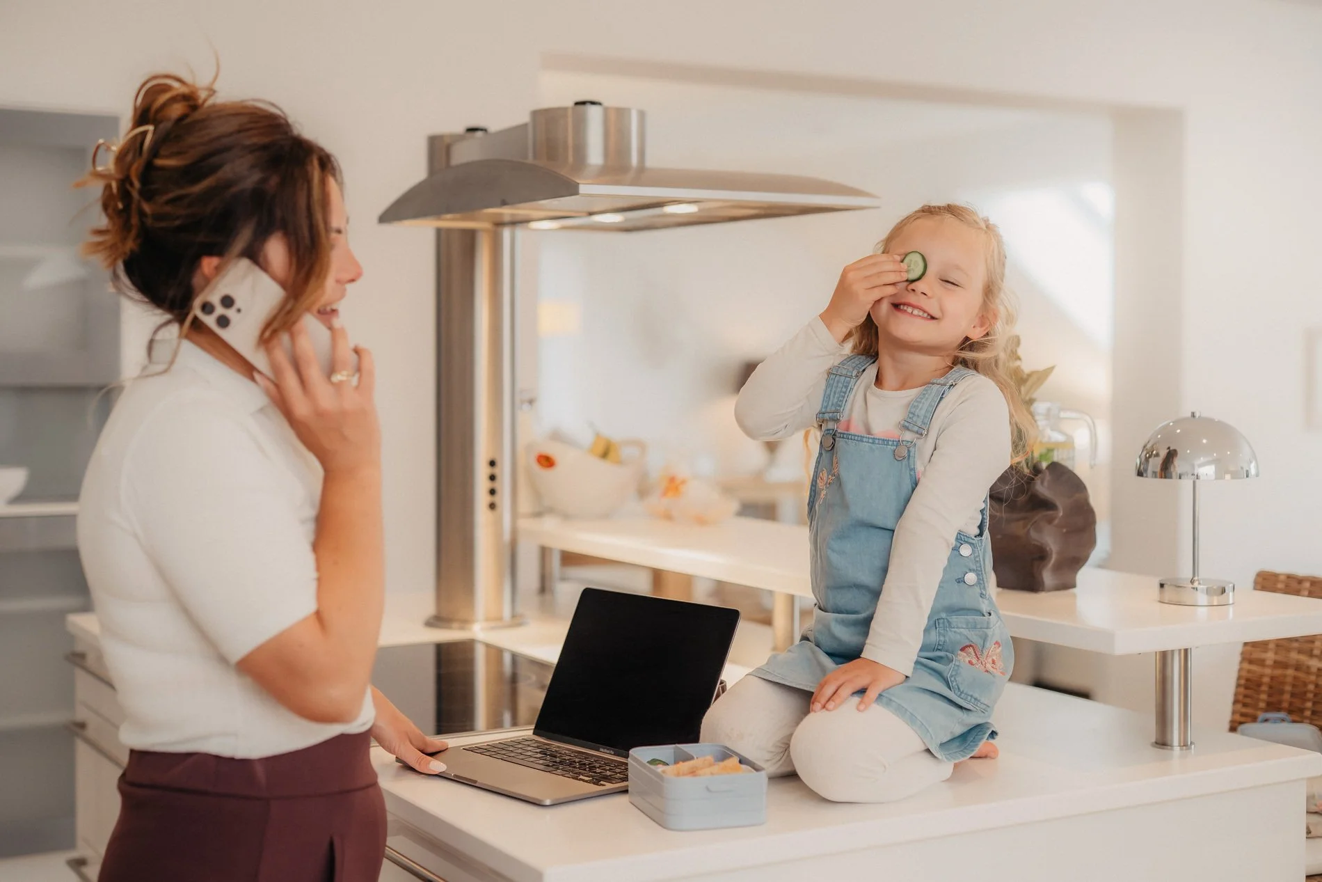 Eine lachende kleine Tochter sitzt auf einer Küchenarbeitsplatte, spielt mit einem Gurkenscheibchen vor einem lachenden Erwachsenen, der mit einem Handy spricht, in einer modernen Küche.