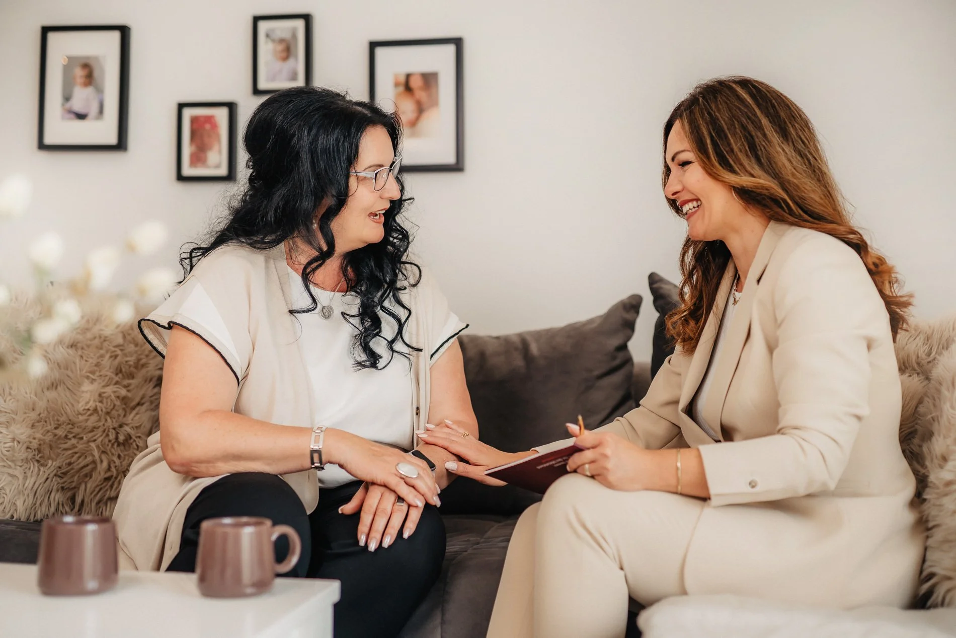 Zwei Frauen sitzen sich gegenüber auf einem Sofa, lächeln und halten die Hände des anderen. Im Hintergrund befinden sich Fotos an der Wand.