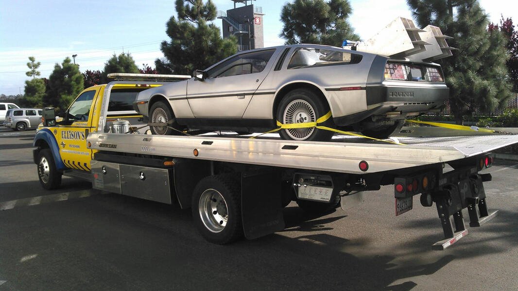 A tow truck carrying a DeLorean DMC-12 in a parking lot with trees in the background.