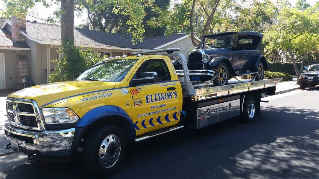 A yellow and blue tow truck carrying a vintage black and blue automobile.