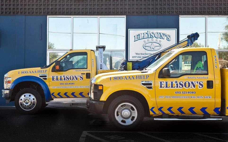 Two yellow tow trucks with blue accents parked in front of a building with large glass windows and a sign for Ellison's, a roadside service company in Montana.