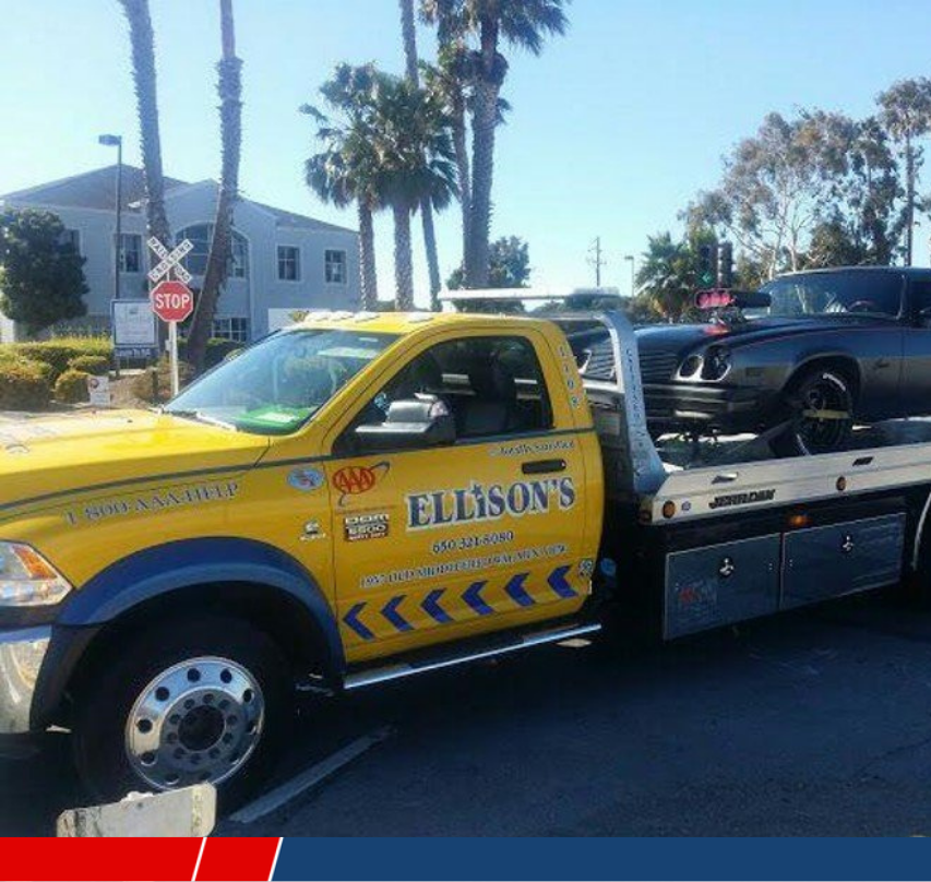 Yellow and blue tow truck carrying a black sports car on its flatbed, parked near a stop sign and building with palm trees nearby.
