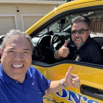 Two men smiling and giving thumbs up, one outside the car and one inside the car, which is yellow.