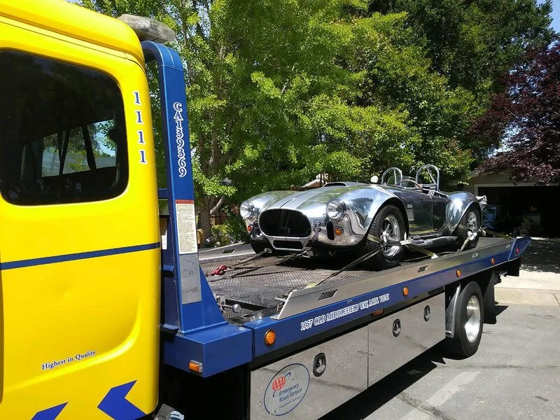 A vintage silver race car on a flatbed tow truck with a yellow cab, parked in a residential area with trees in the background.