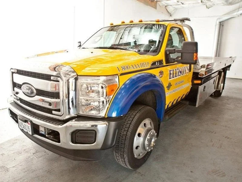 Yellow and blue ambulance tow truck with company name Ellisons and phone number 1-800-4-AM-HELP on the side, parked indoors.