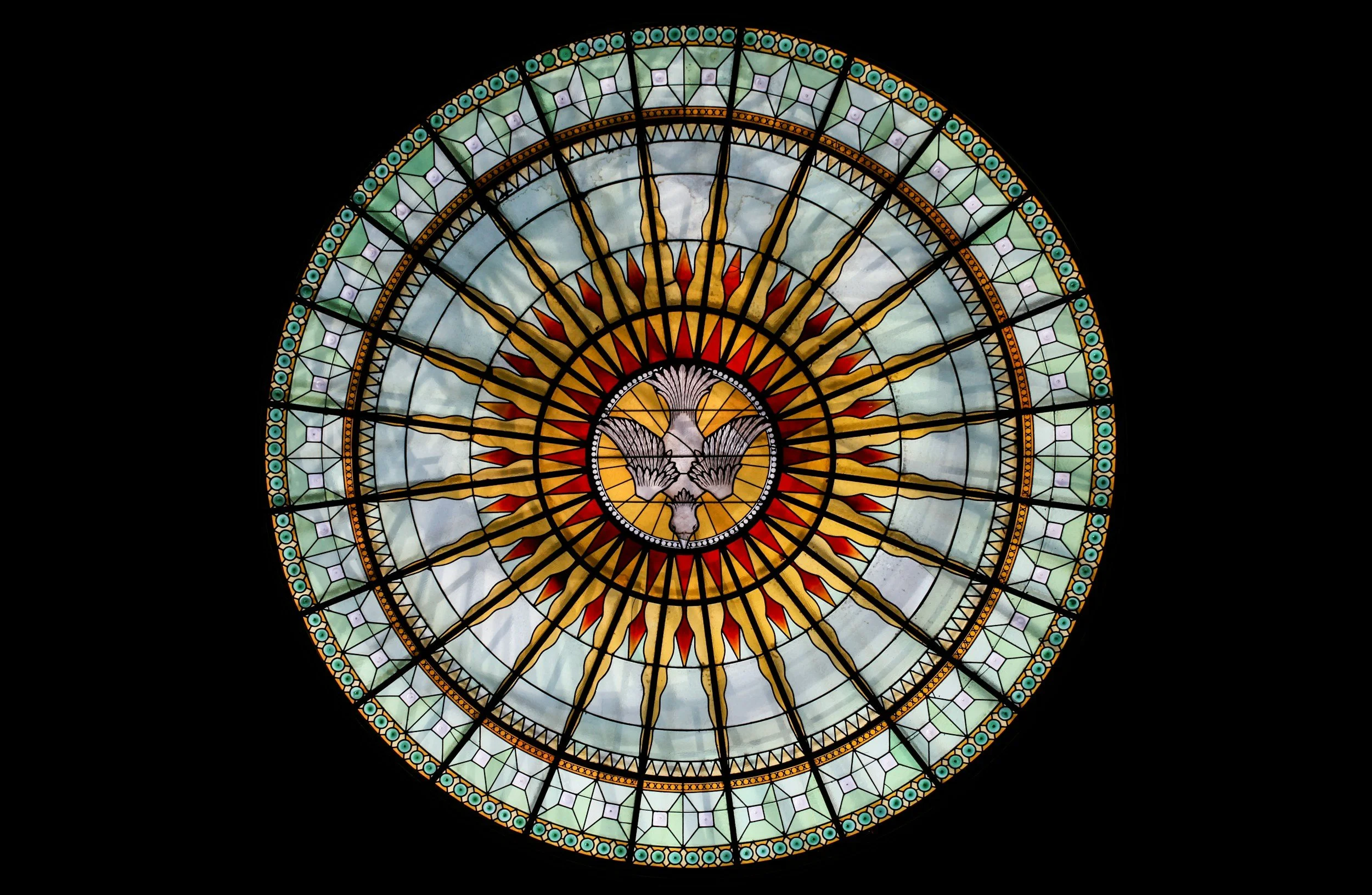 A circular stained glass window featuring a dove at the center, with rays extending outward, set against a cloudy sky background.