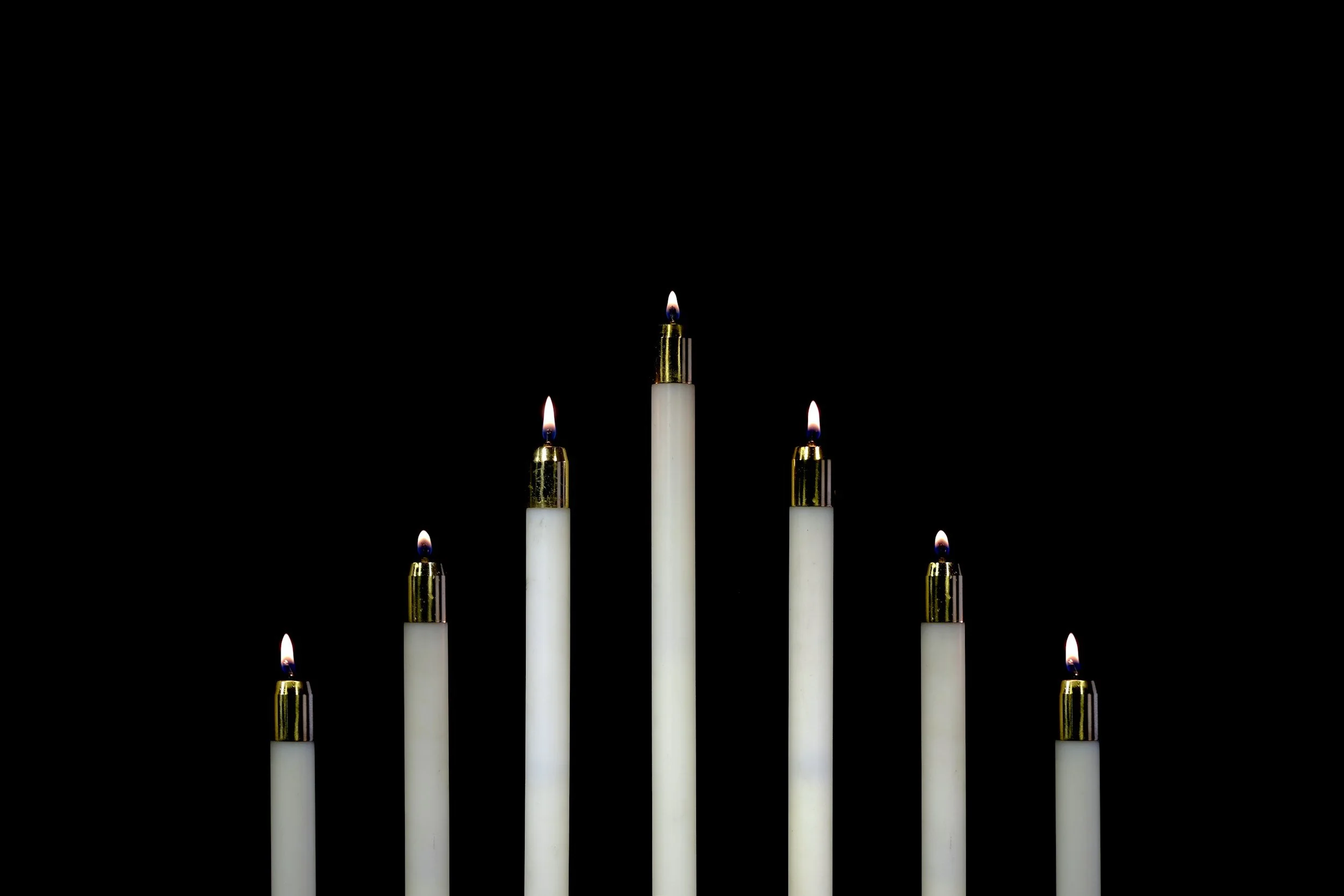 Six white candles with gold holders burning against a black background.