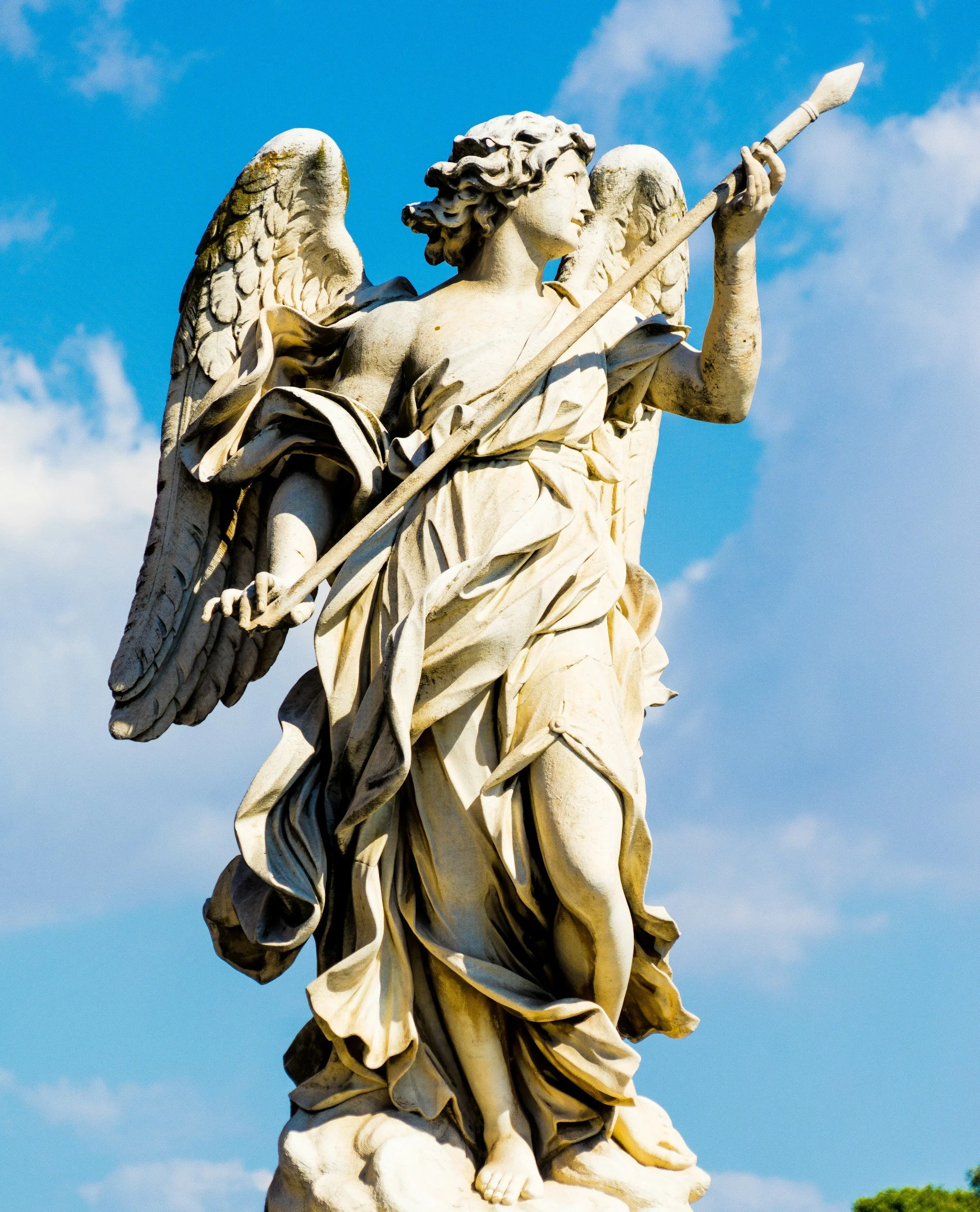 Close-up of a white marble angel sculpture holding a spear, with detailed wings and flowing robes, against a bright blue sky with some clouds.