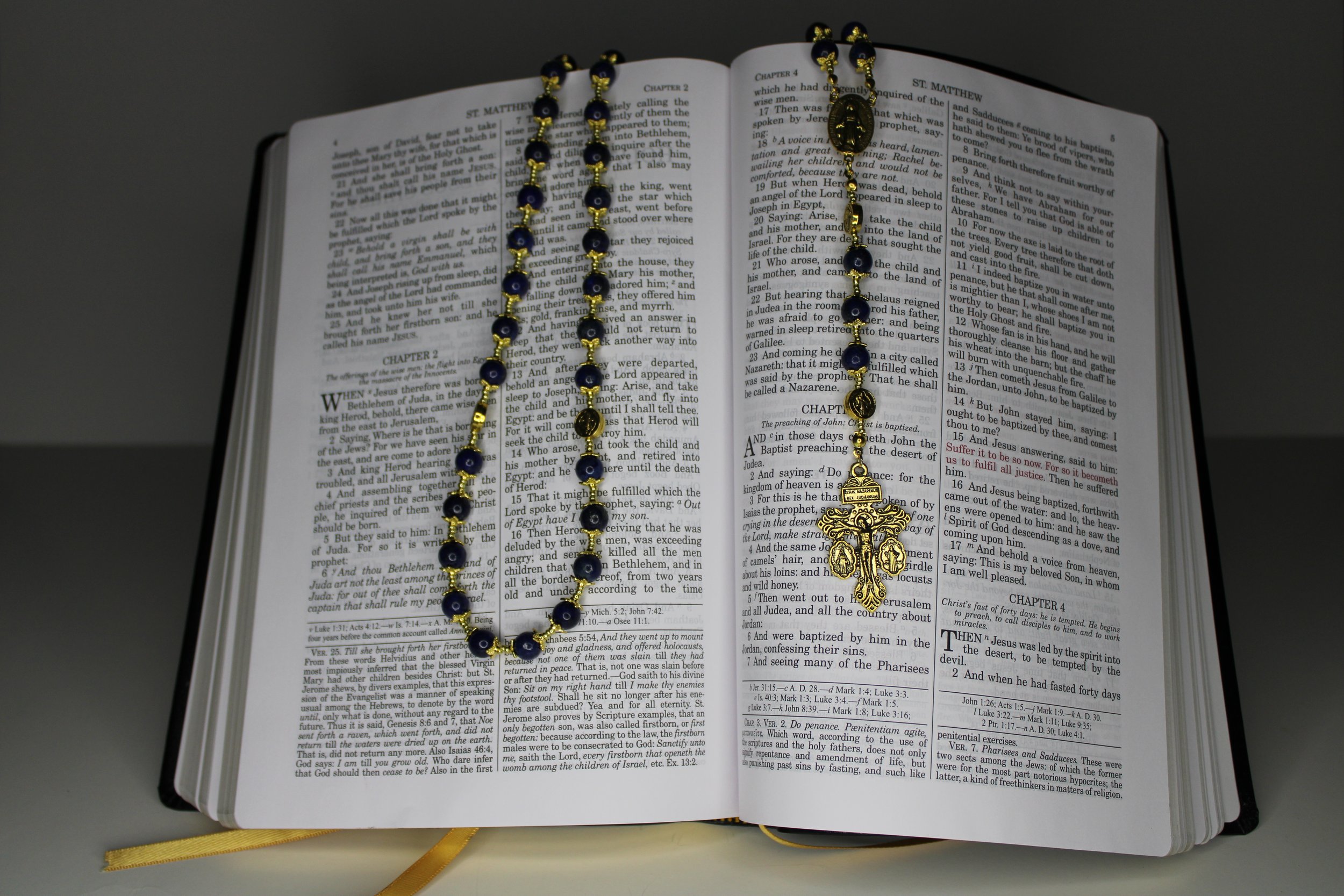 An open Bible with a blue and gold prayer rosary laid across its pages, featuring a gold cross pendant.