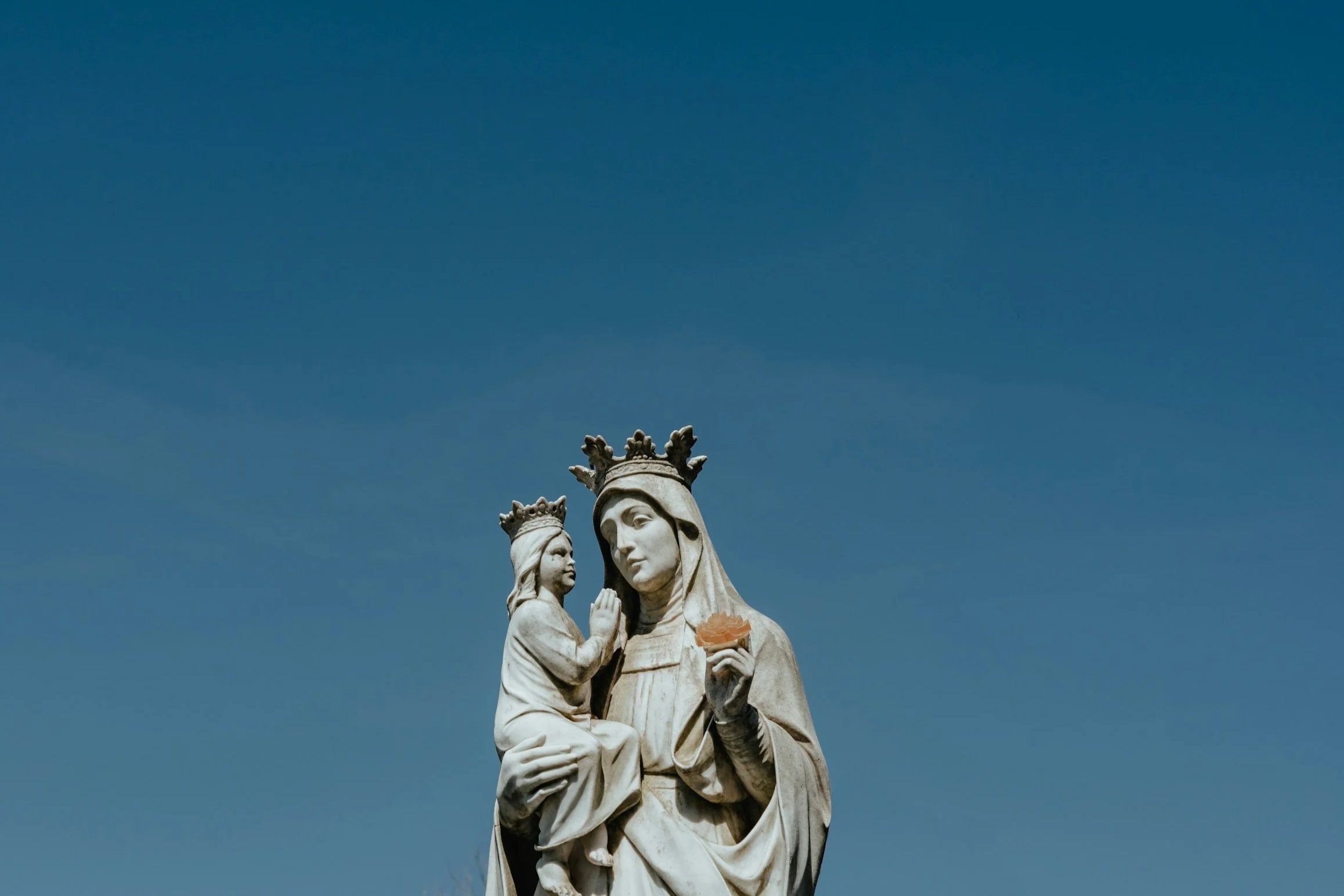 Statue of the Virgin Mary holding Baby Jesus, with two crowns on her head, against a blue sky.