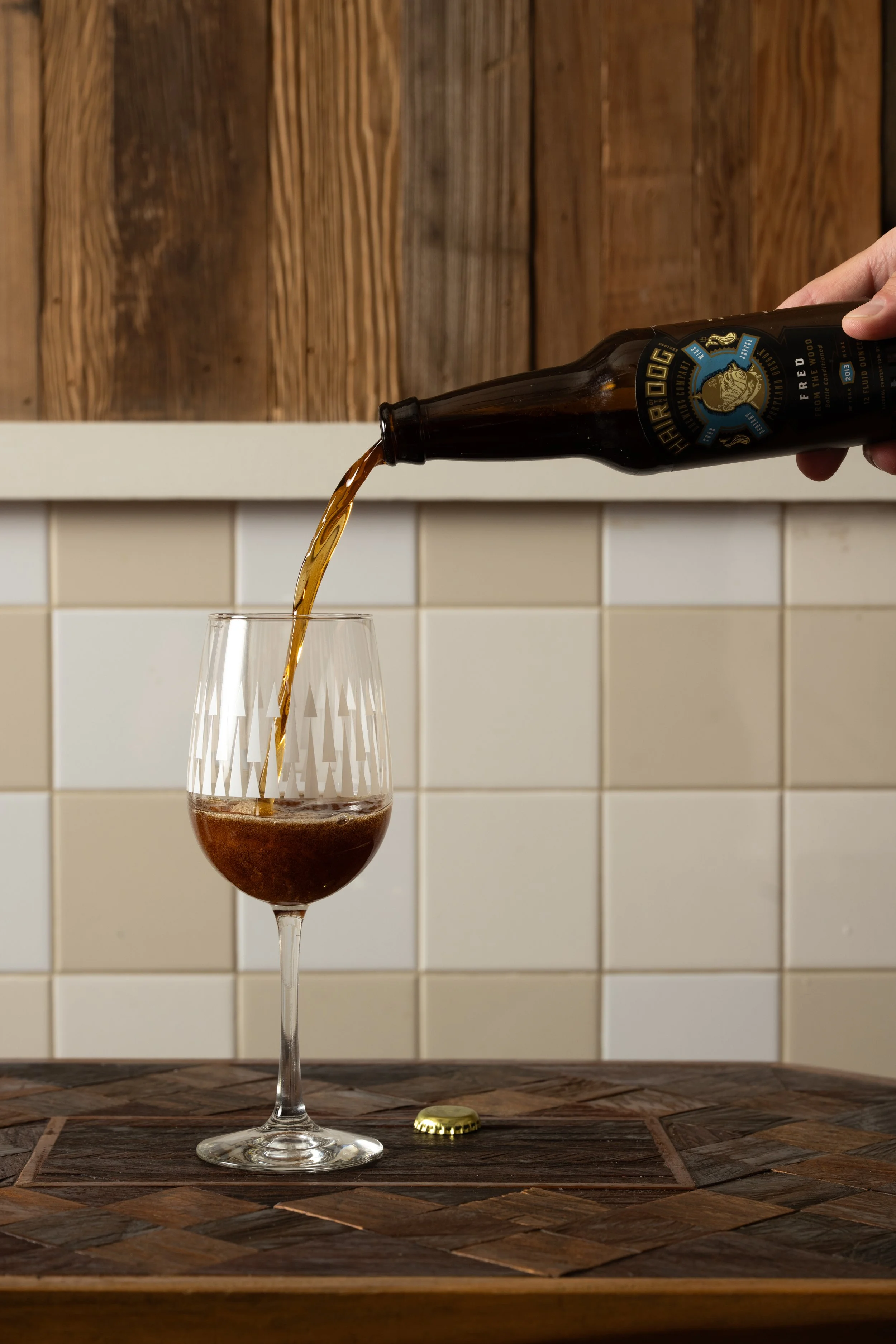 Beer being poured from a bottle into a patterned Tørst glass on a wood-inlay table against white tile and wood panel walls