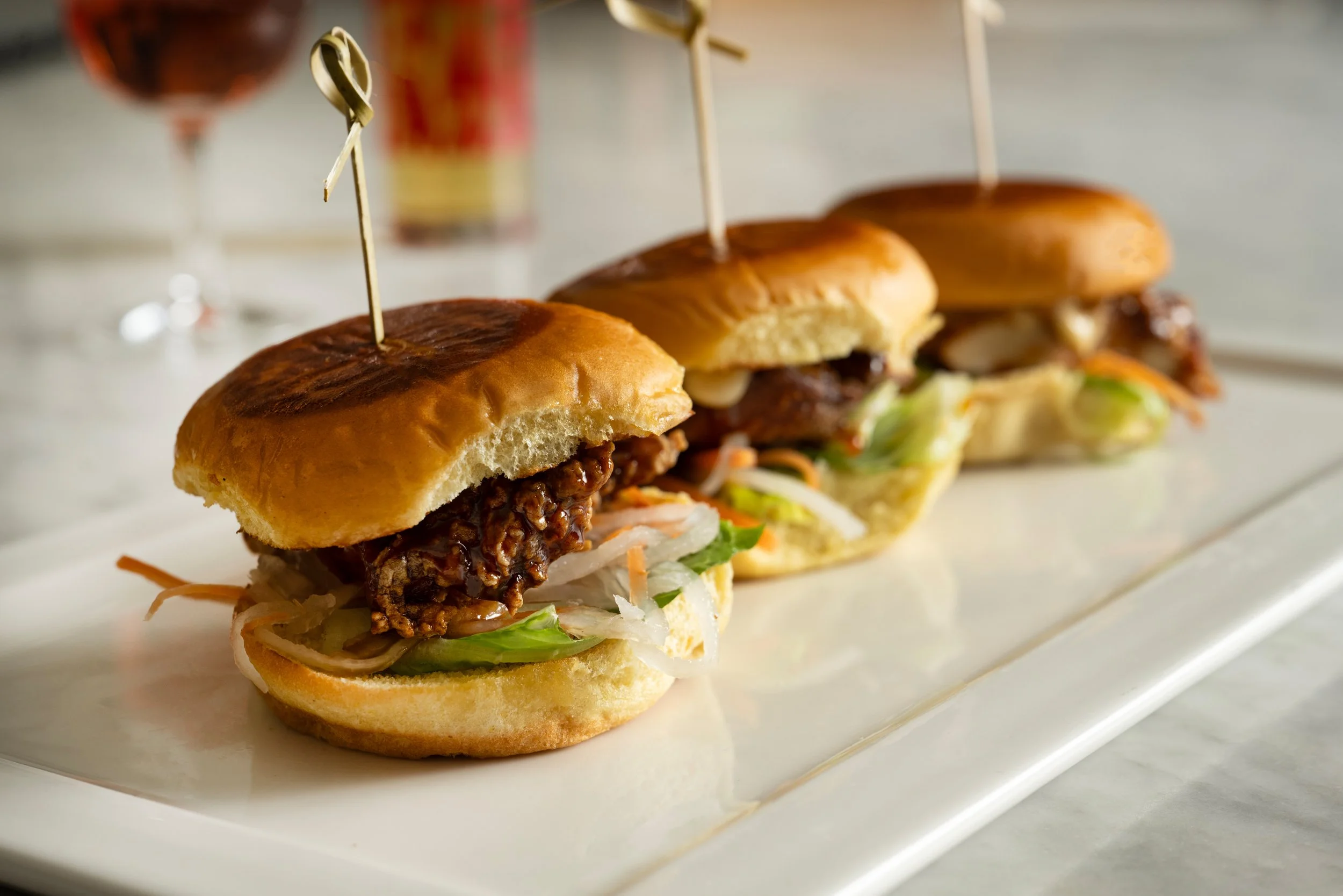 Three fried chicken sandwich sliders with chili miso honey glaze, pickled carrot and daikon, and garlic aioli on brioche buns, served on a marble board