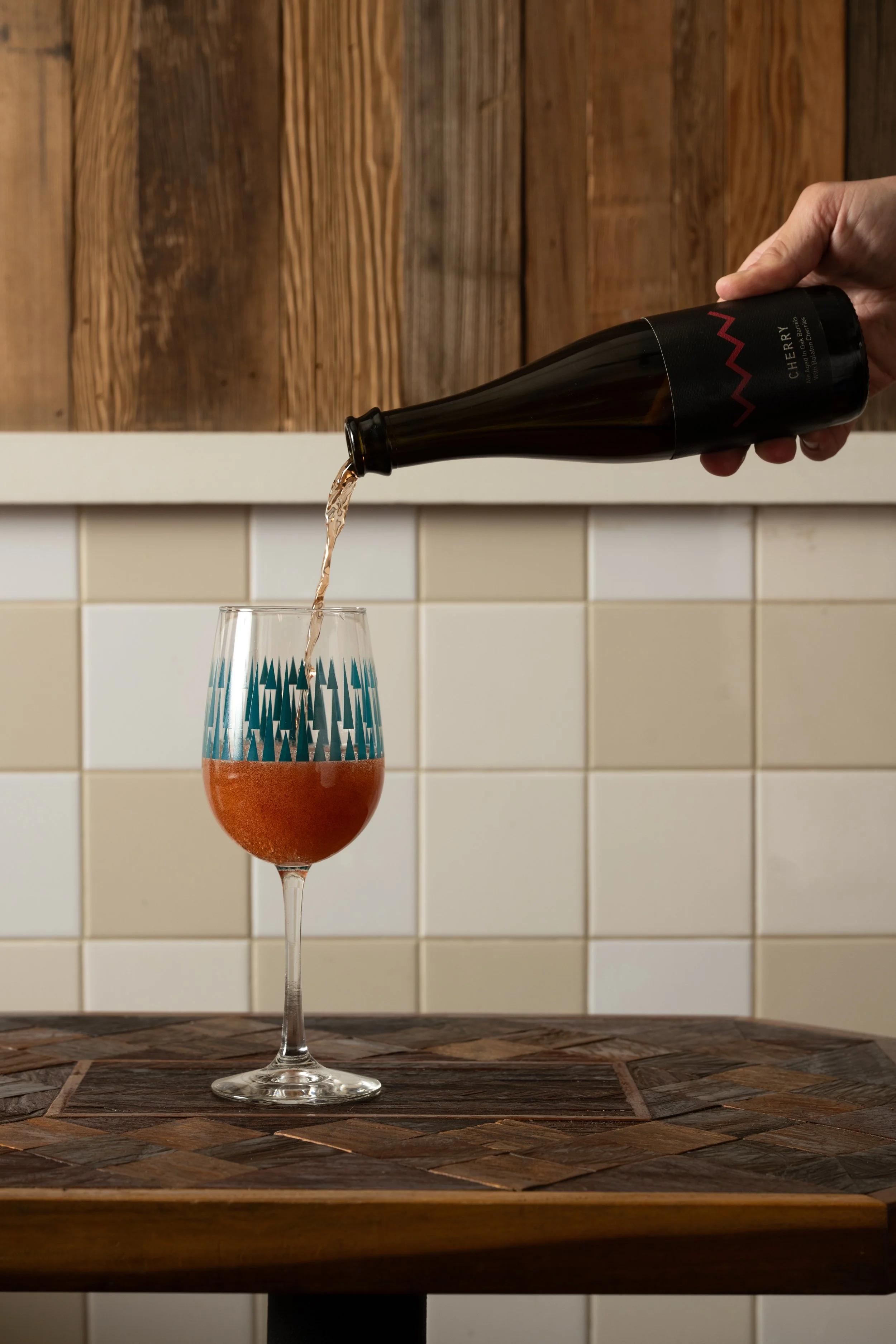 Beer being poured from a bottle into a teal patterned Tørst glass on a wood-inlay table against white tile and wood panel walls