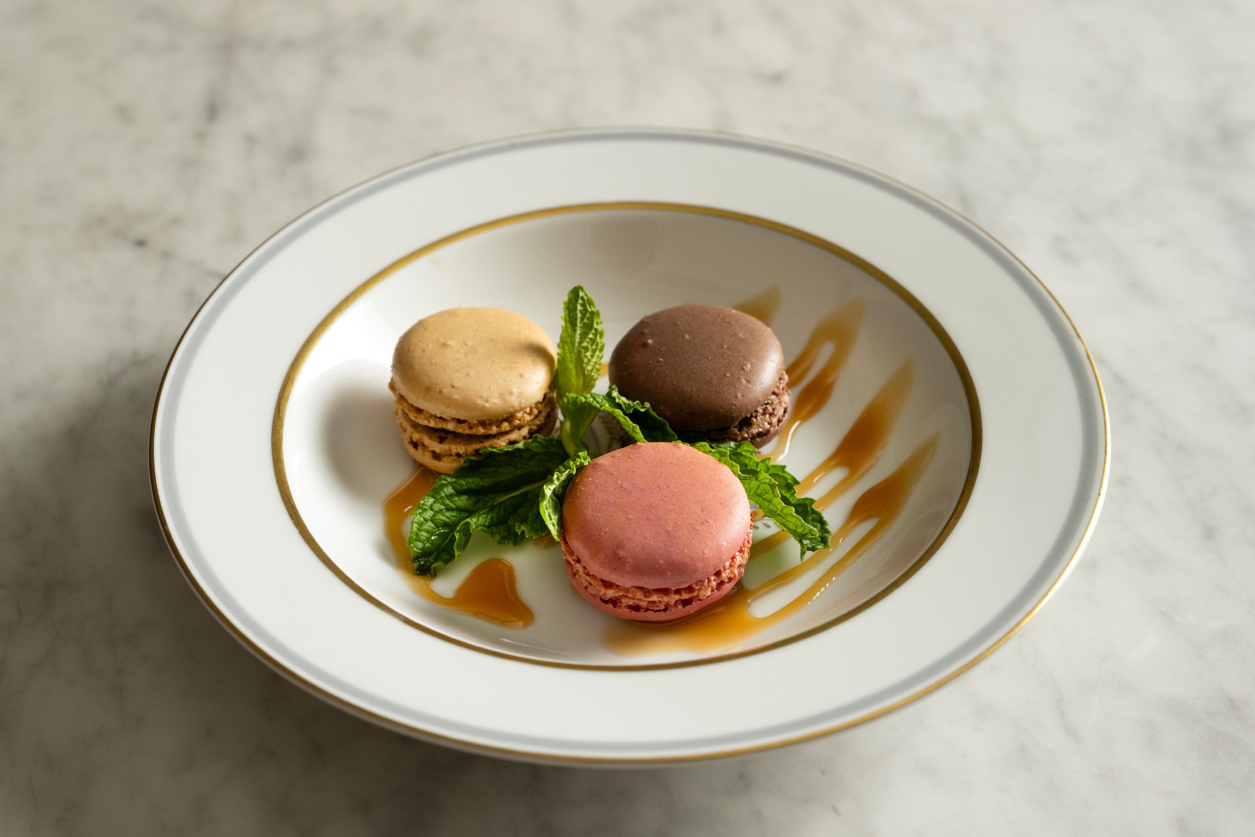 Three mini macarons in coffee, chocolate, and raspberry arranged on a gold-rimmed plate with fresh mint and caramel drizzle