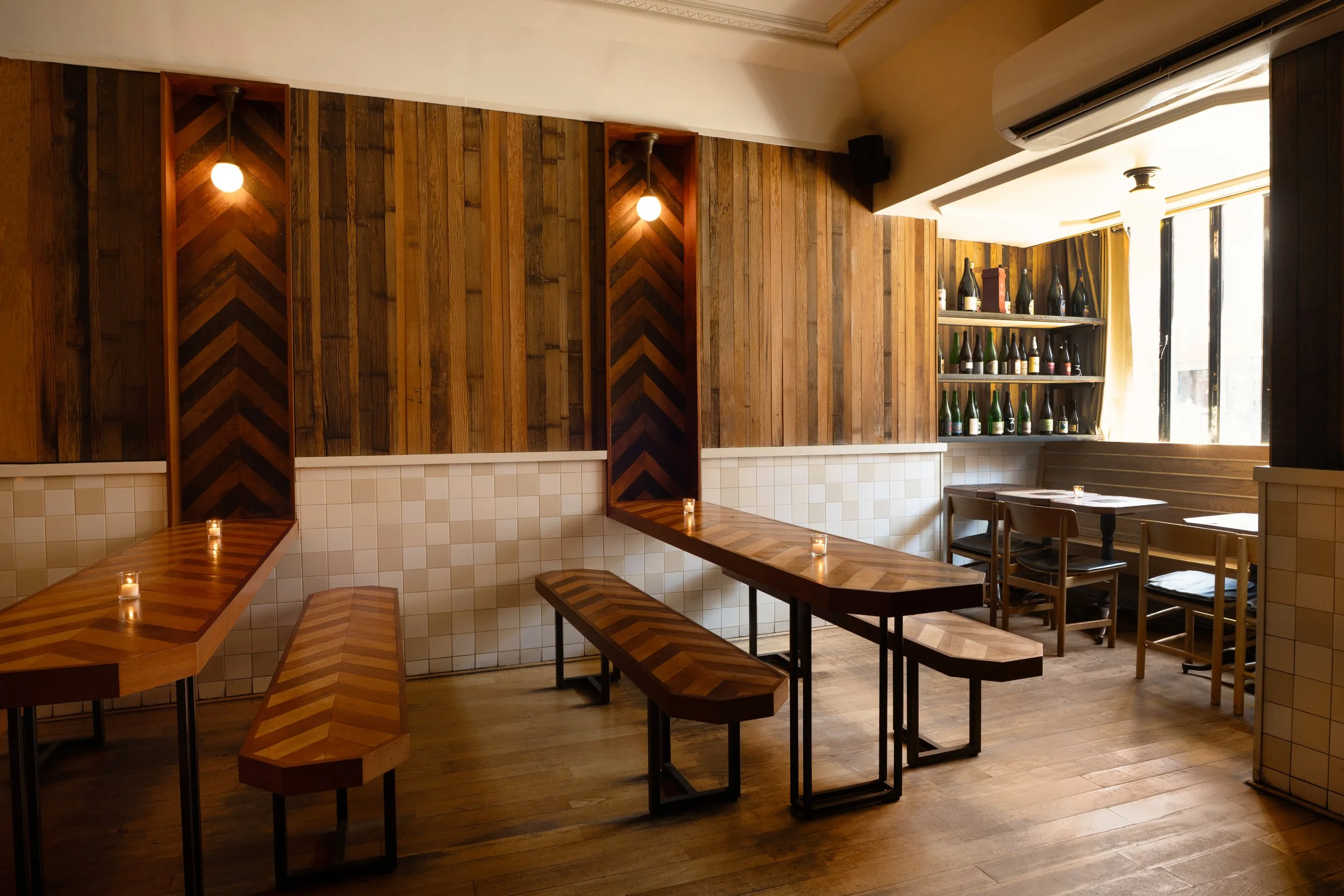 Warm, candlelit dining room at Tørst with herringbone wood walls, long communal tables, and shelves of bottled beer by the window