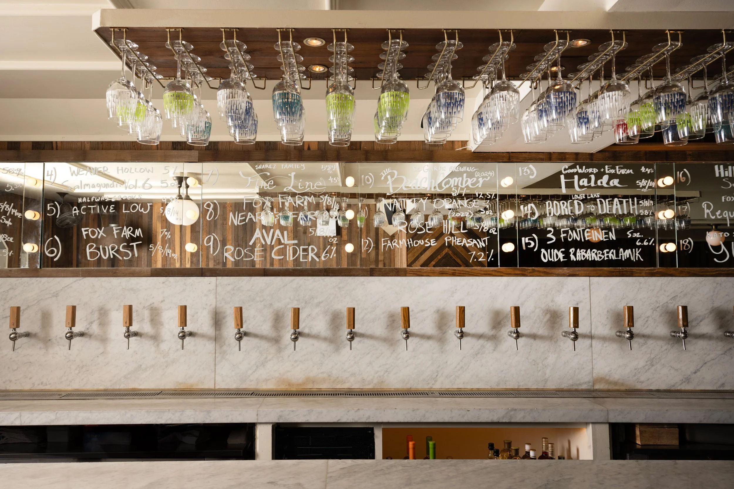 Close-up of Tørst's draft tap wall with handwritten beer list on mirrored backbar and colorful patterned glassware hanging overhead