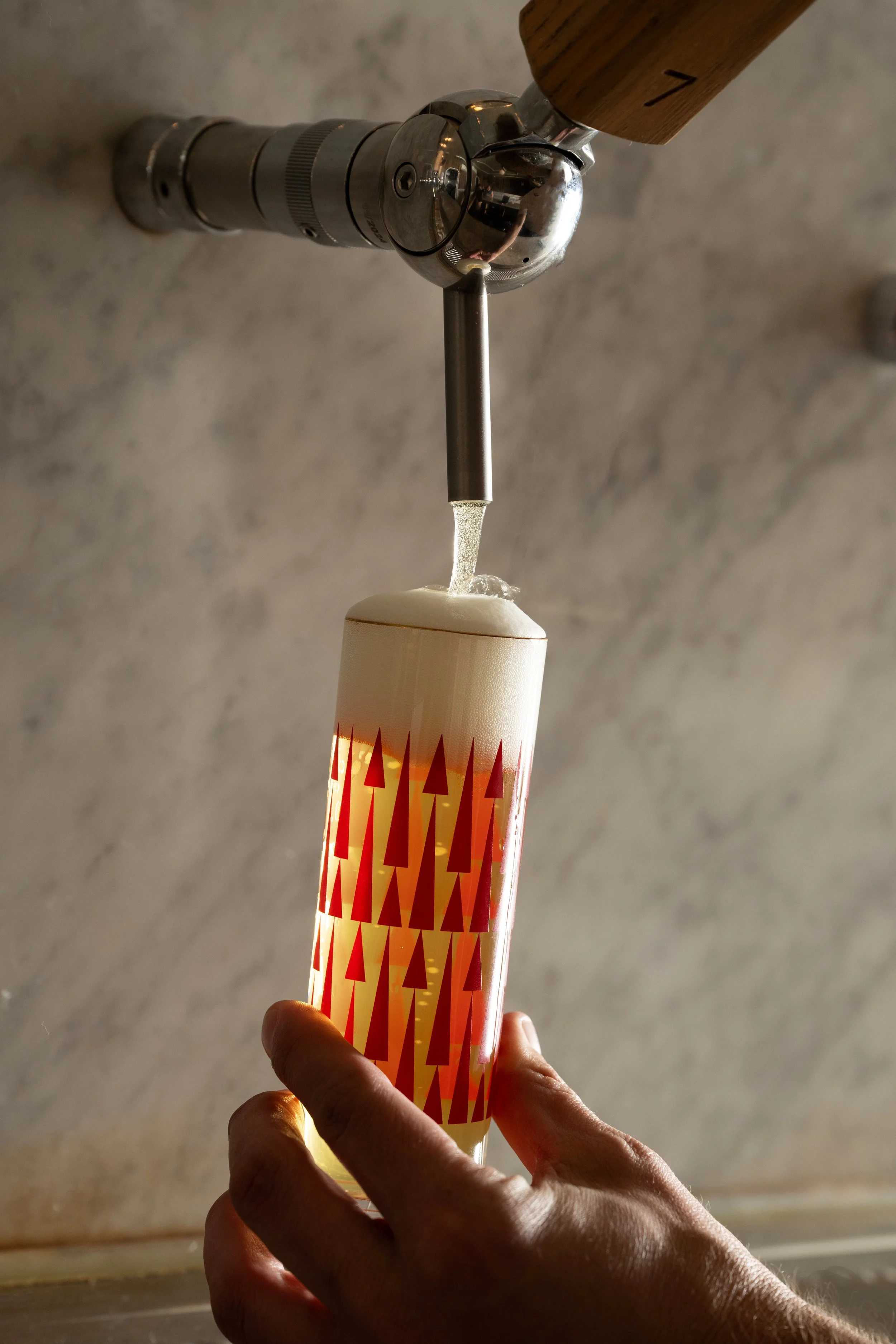Freshly poured red patterned Tørst pilsner glass filled with golden beer and a tight head of foam, resting on the marble bar