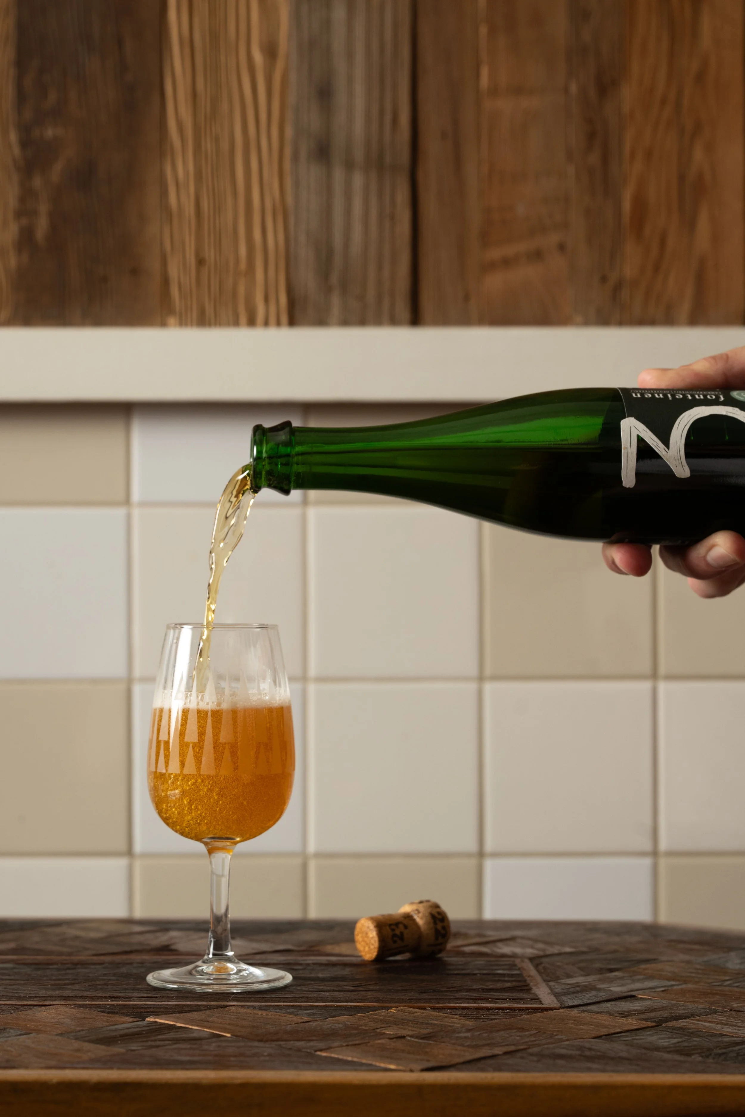 Bottle of beer being poured into a glass on a wood-inlay table, cork resting beside it