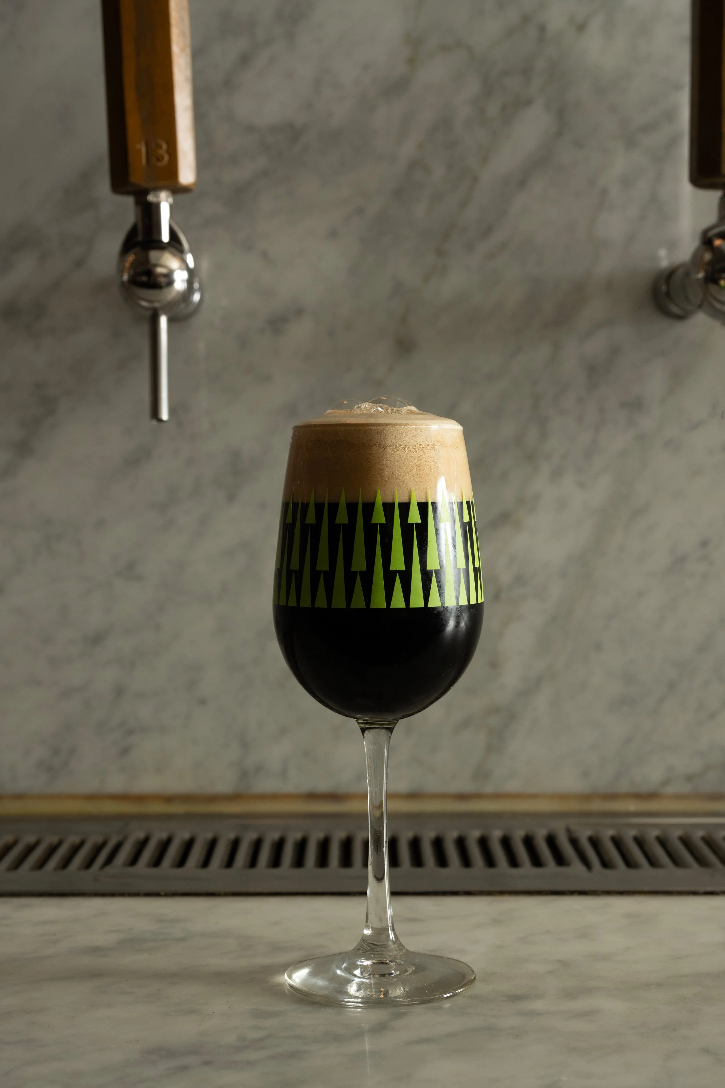 Dark stout with a tan foam head in a green patterned Tørst glass resting on the marble bar with two wooden tap handles visible