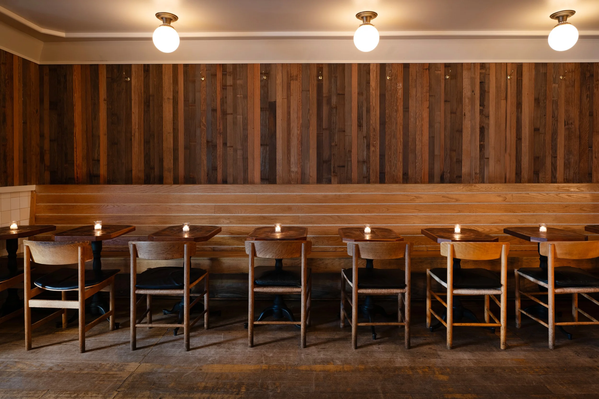 Row of wooden chairs and small tables along a banquette at Tørst, lit by globe pendant lights