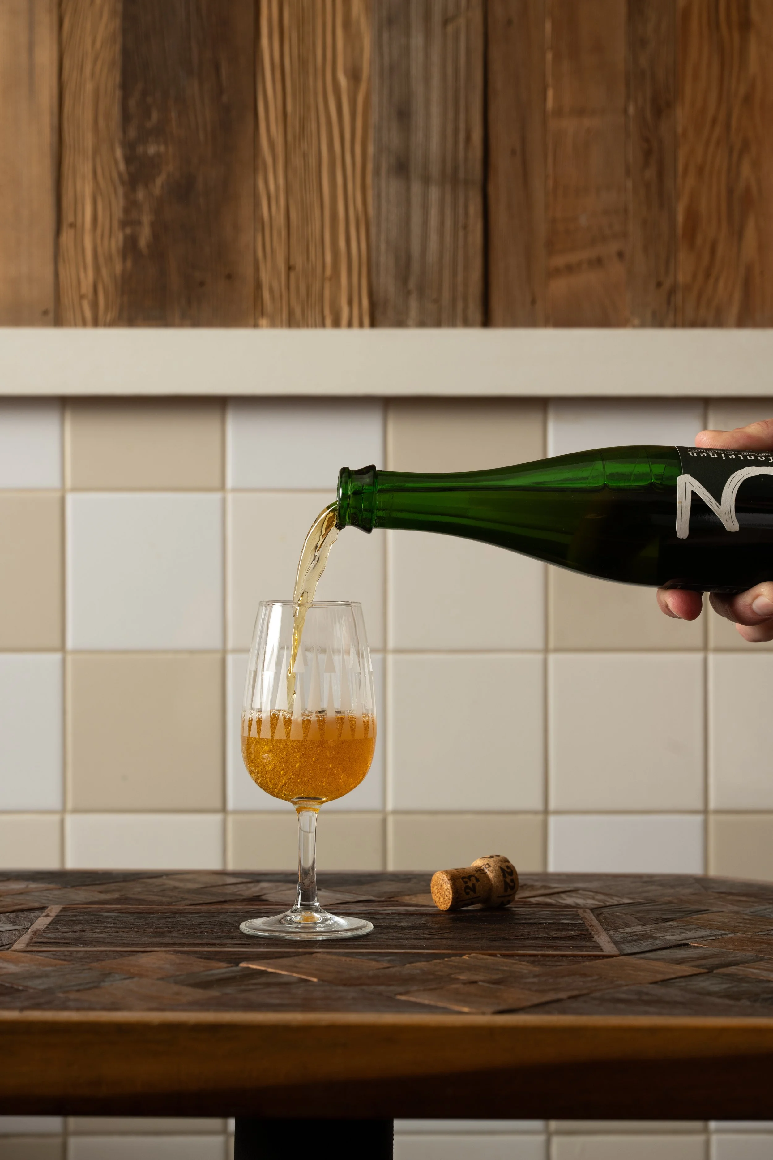 Bottle of beer being poured into a glass on a wood-inlay table, cork resting beside it