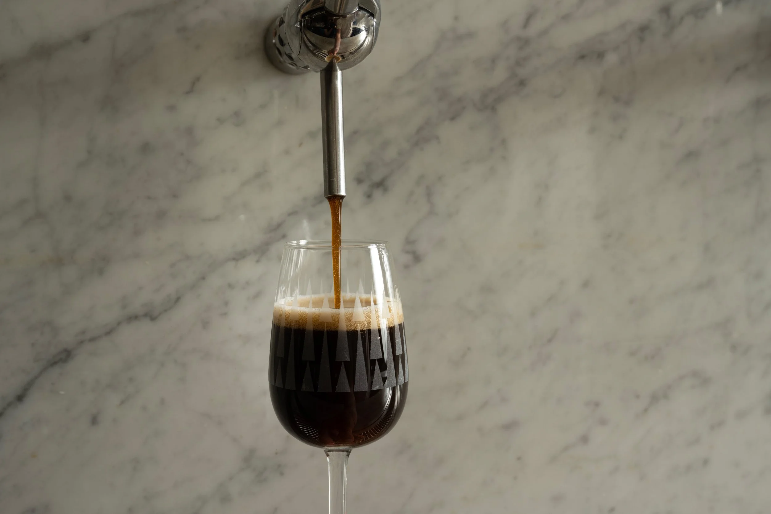 Dark beer being poured from a draft tap into a clear patterned Tørst glass against a marble bar back
