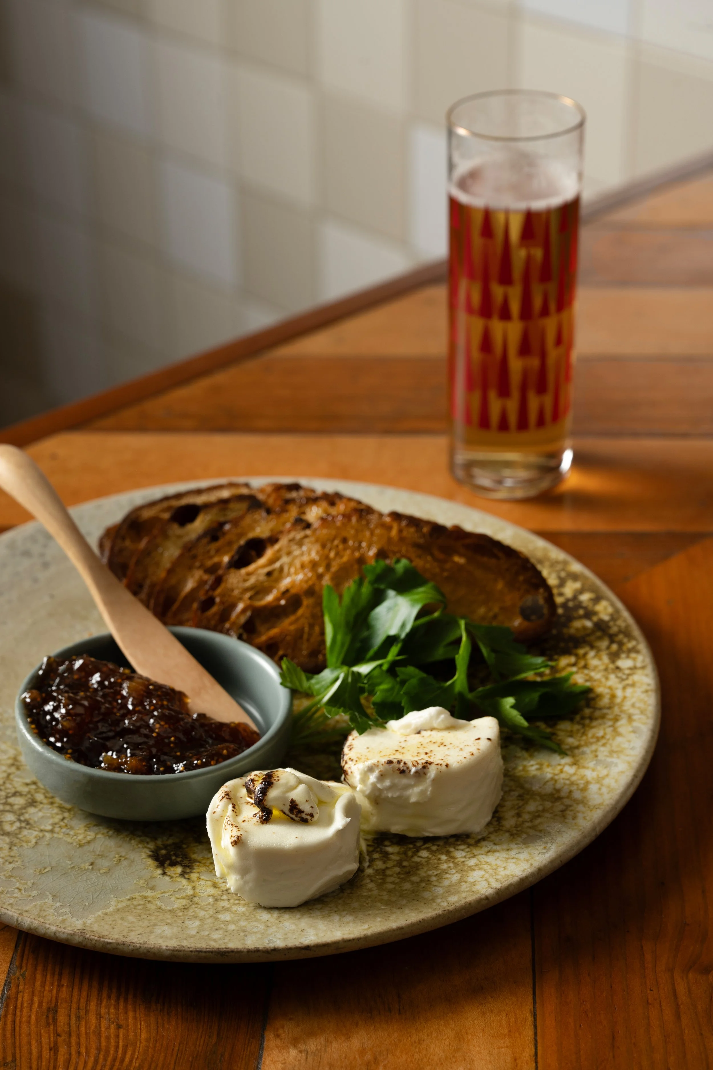 Cheese plate with soft cheese, seasonal jam, fresh herbs, and sourdough crostinis served alongside a patterned Tørst beer glass