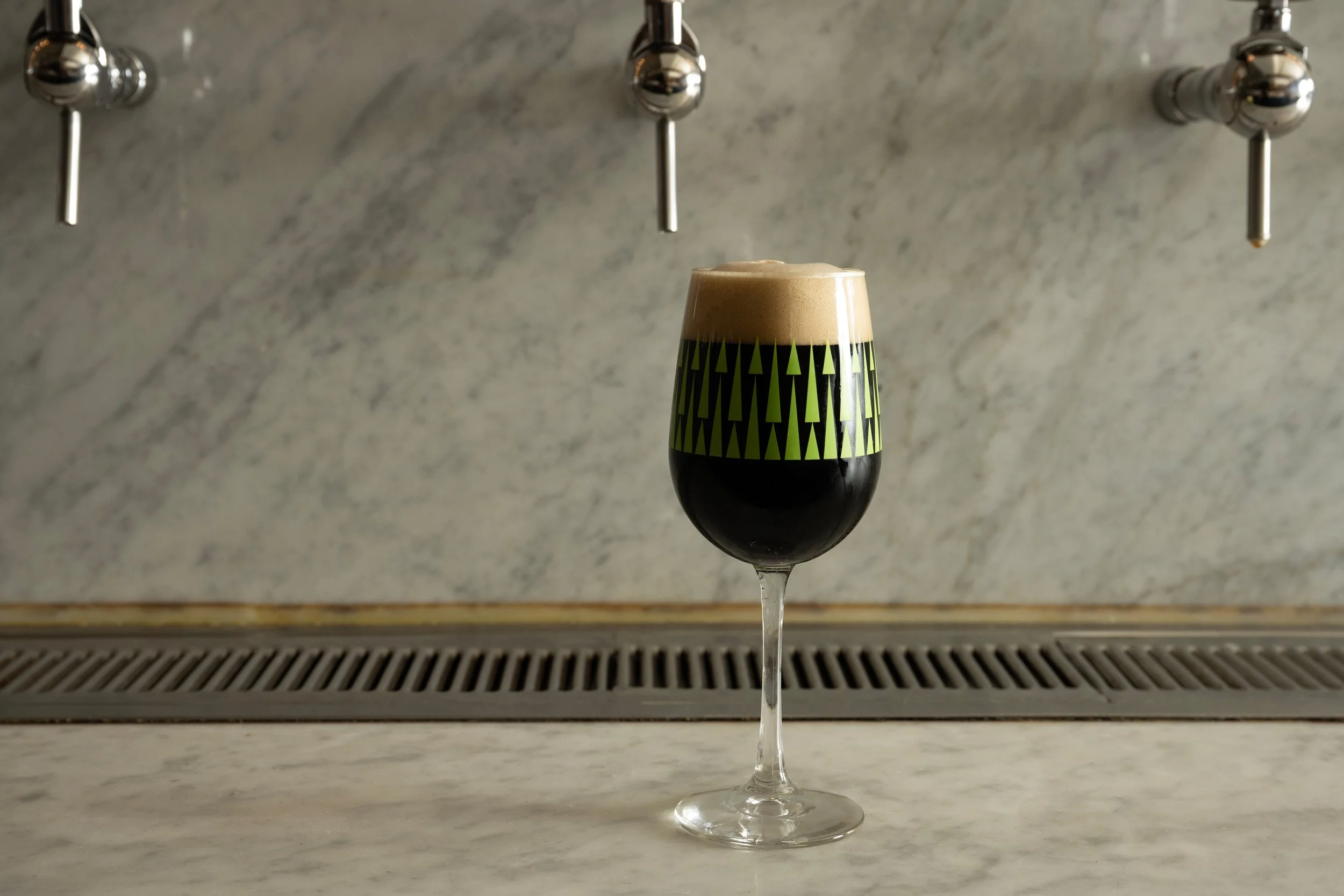 Dark stout with a tan foam head in a green patterned Tørst glass resting on the marble bar with draft tap handles visible above