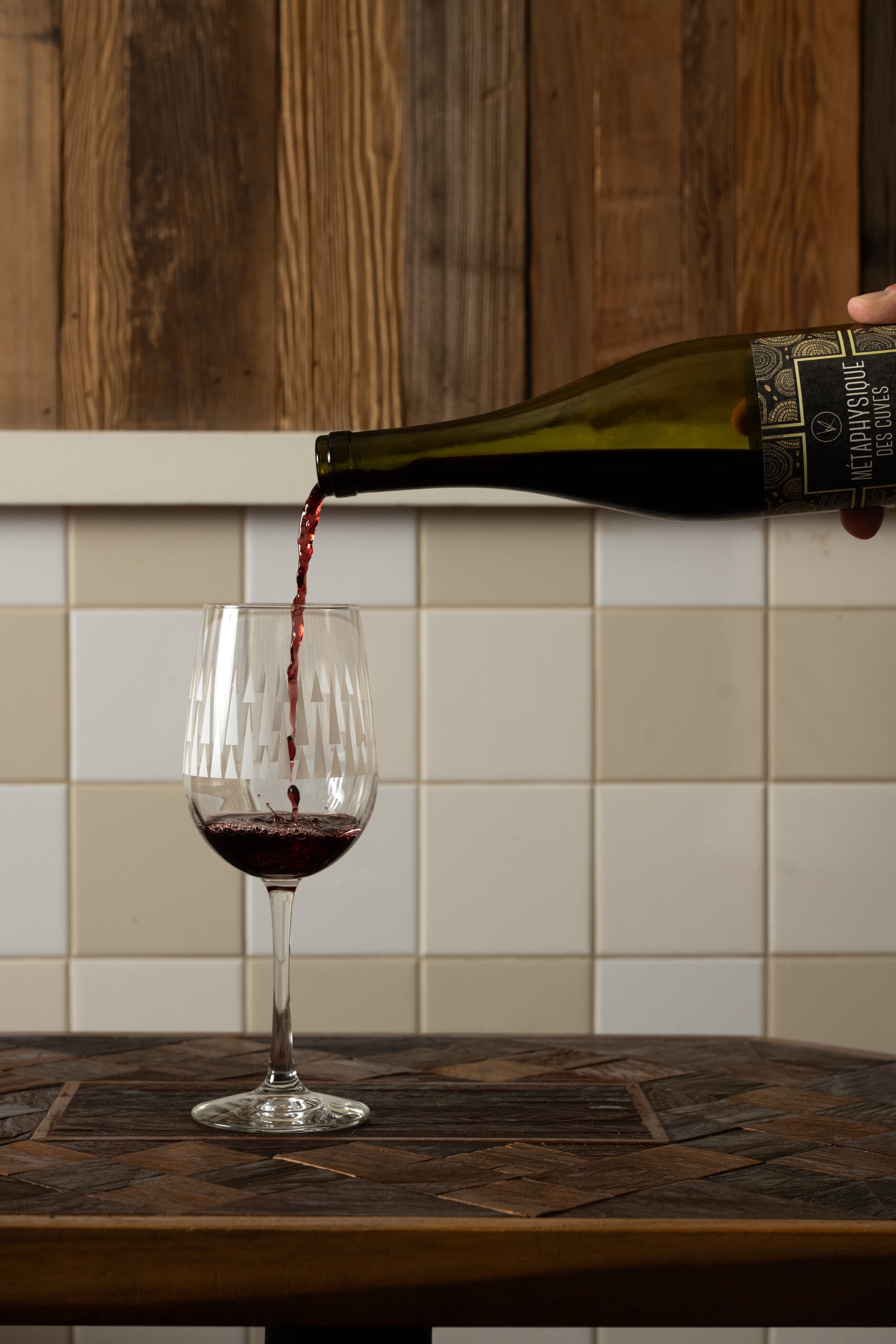 Red wine being poured from a bottle into a clear patterned Tørst glass on a wood-inlay table against white tile and wood panel walls