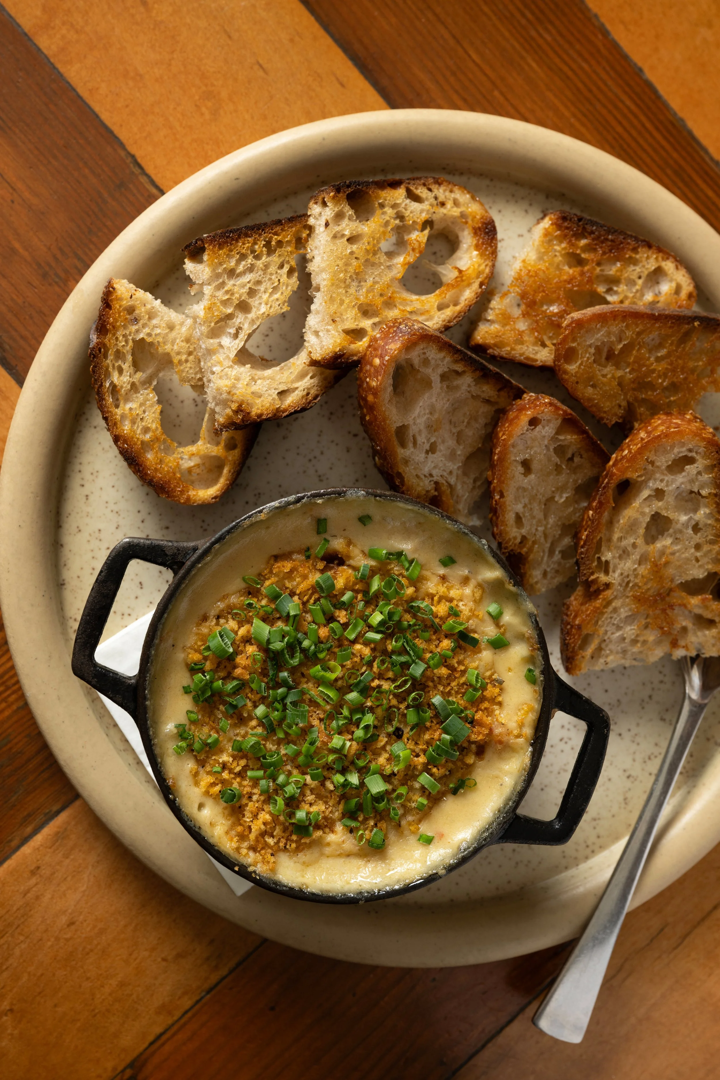 Hot crab dip topped with breadcrumbs and chives in a cast iron ramekin, served with toasted sourdough crostinis
