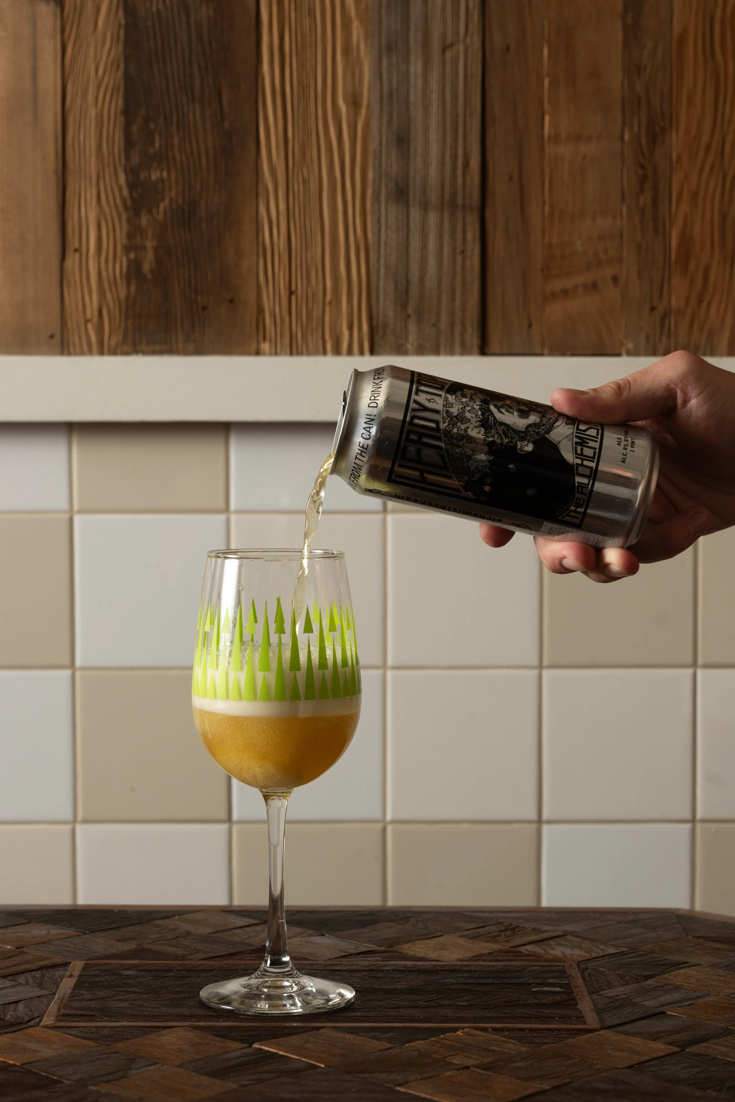Can of beer being poured from a can into a green patterned Tørst glass on a wood-inlay table against white tile and wood panel walls
