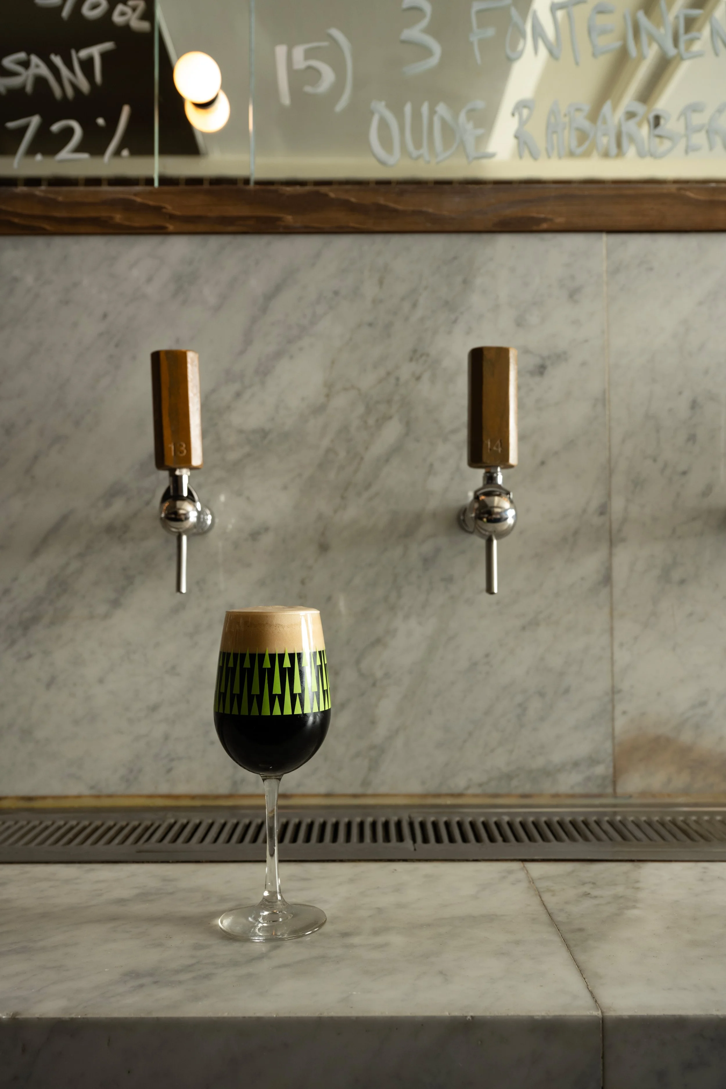 Dark stout in a green patterned Tørst glass sitting on the marble bar in front of wooden tap handles 13 and 14, with the handwritten beer list reflected in the mirror above