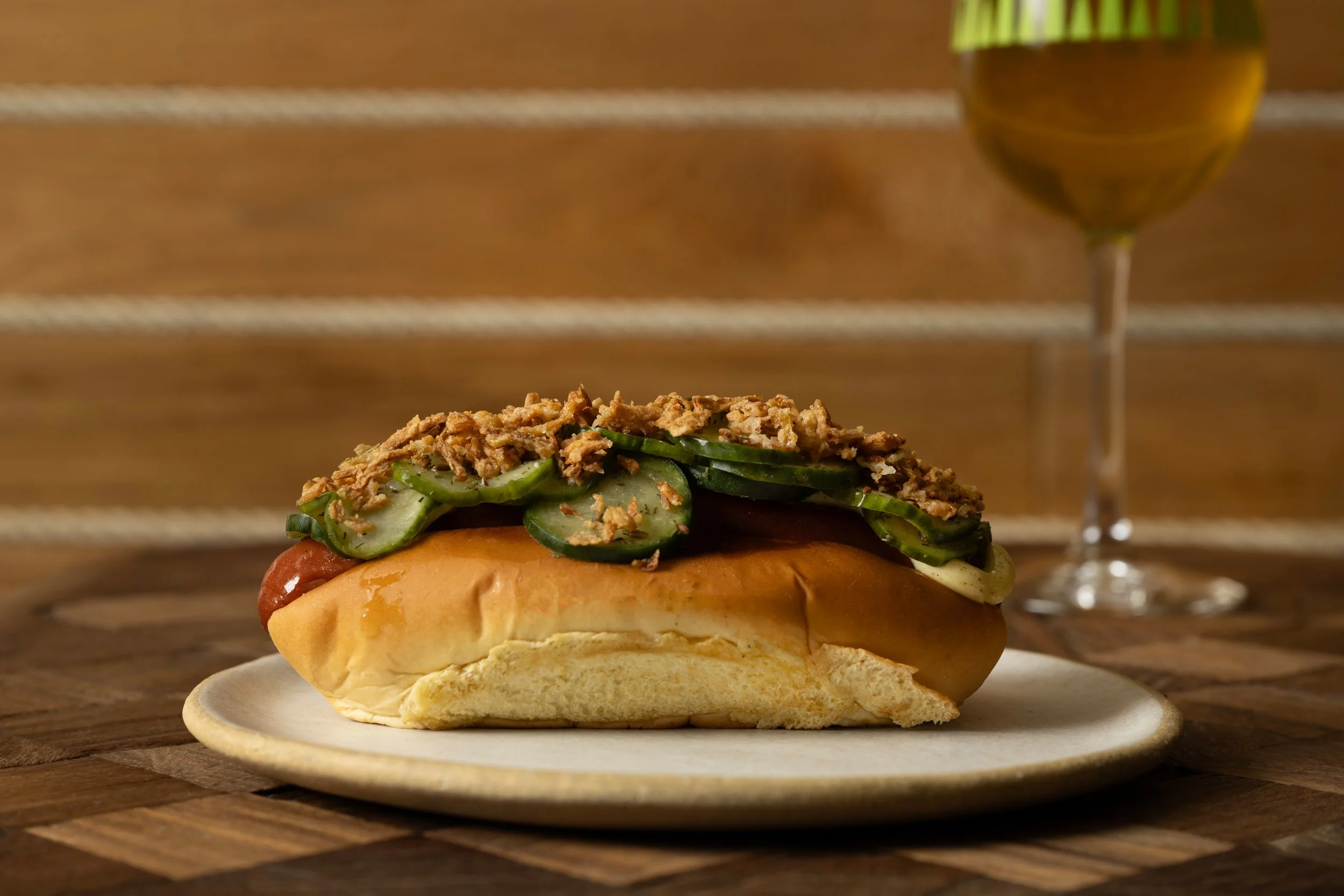 Danish beef dog with curry remoulade, quick-pickled cucumbers, and fried onions on a brioche bun, served alongside a patterned Tørst beer glass